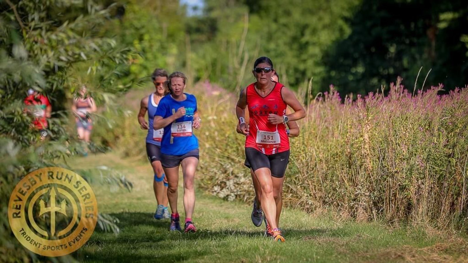A group of runners is participating in an outdoor race on a grassy trail surrounded by tall wildflowers. The runners, wearing athletic gear, are in mid-stride. The logo on the bottom left corner reads "Revesby Run Series, Trident Sports Events.