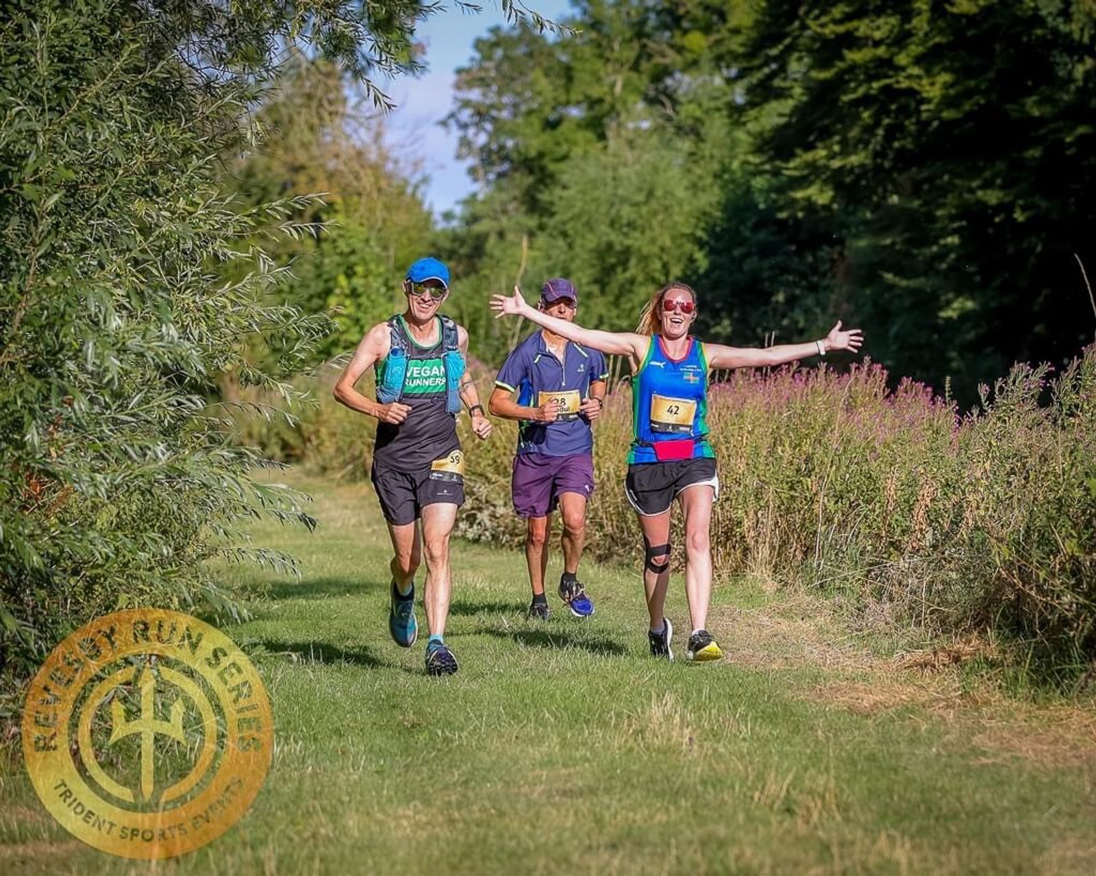 Three runners are on a grassy path surrounded by green foliage during an outdoor race. The runner on the right, wearing a blue and yellow top, has arms outstretched and is smiling at the camera. The runner on the left wears a green top, and the middle runner is in purple. The "Run Belvoir" logo is in the bottom left corner.
