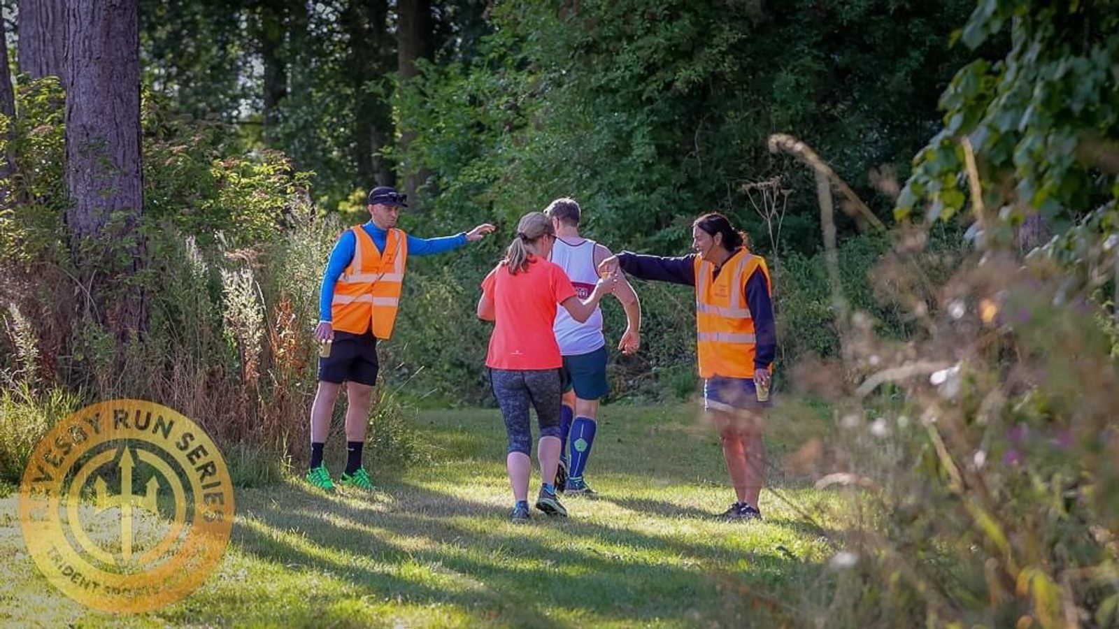 Four people are standing on a grassy path surrounded by trees. Two are wearing orange safety vests, seemingly giving directions or instructions. Two others, in athletic wear, appear to be participants in an event, possibly a run. A logo for the East Run Series is visible in the corner.