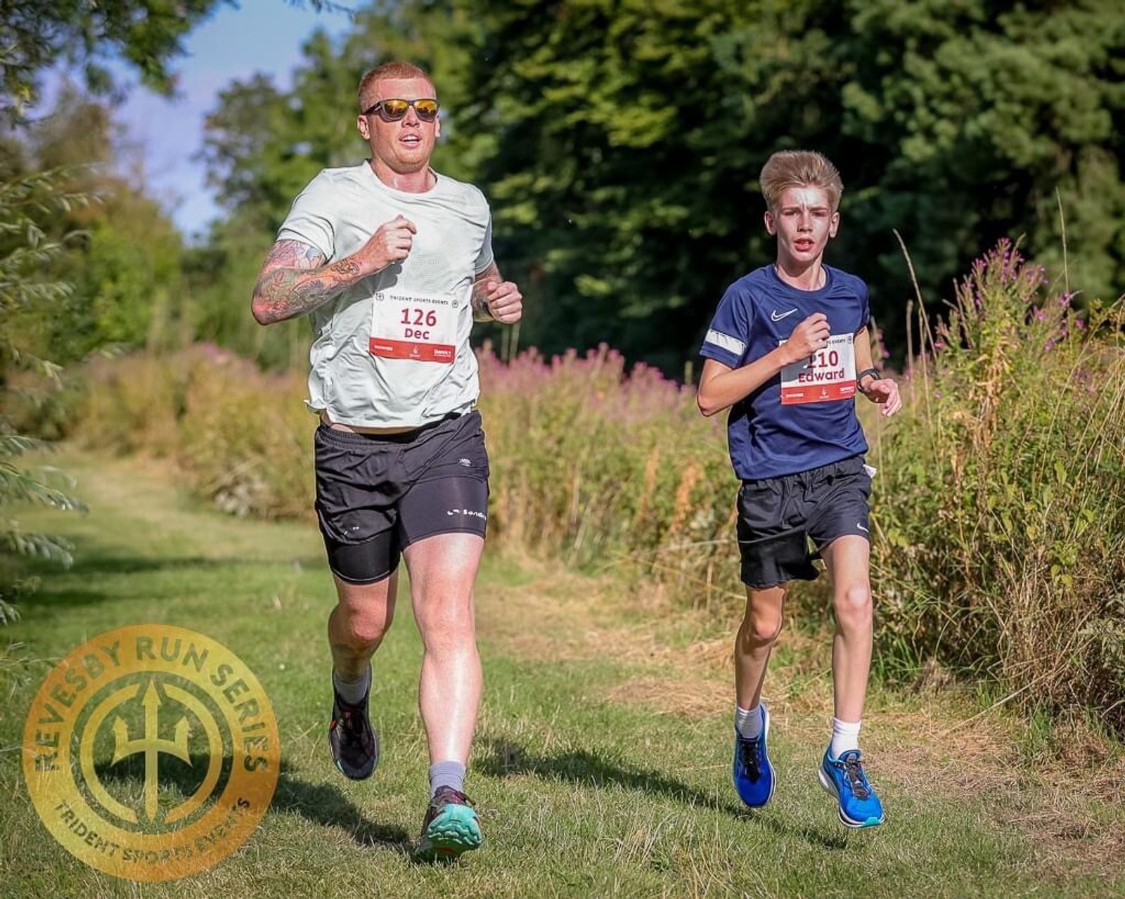 Two runners participating in the "Sevensy Run Series." The adult runner, wearing sunglasses, a light grey shirt, and black shorts, is ahead. The young runner wears a navy blue shirt and shorts. Both have race bibs as they run on a scenic path surrounded by greenery.