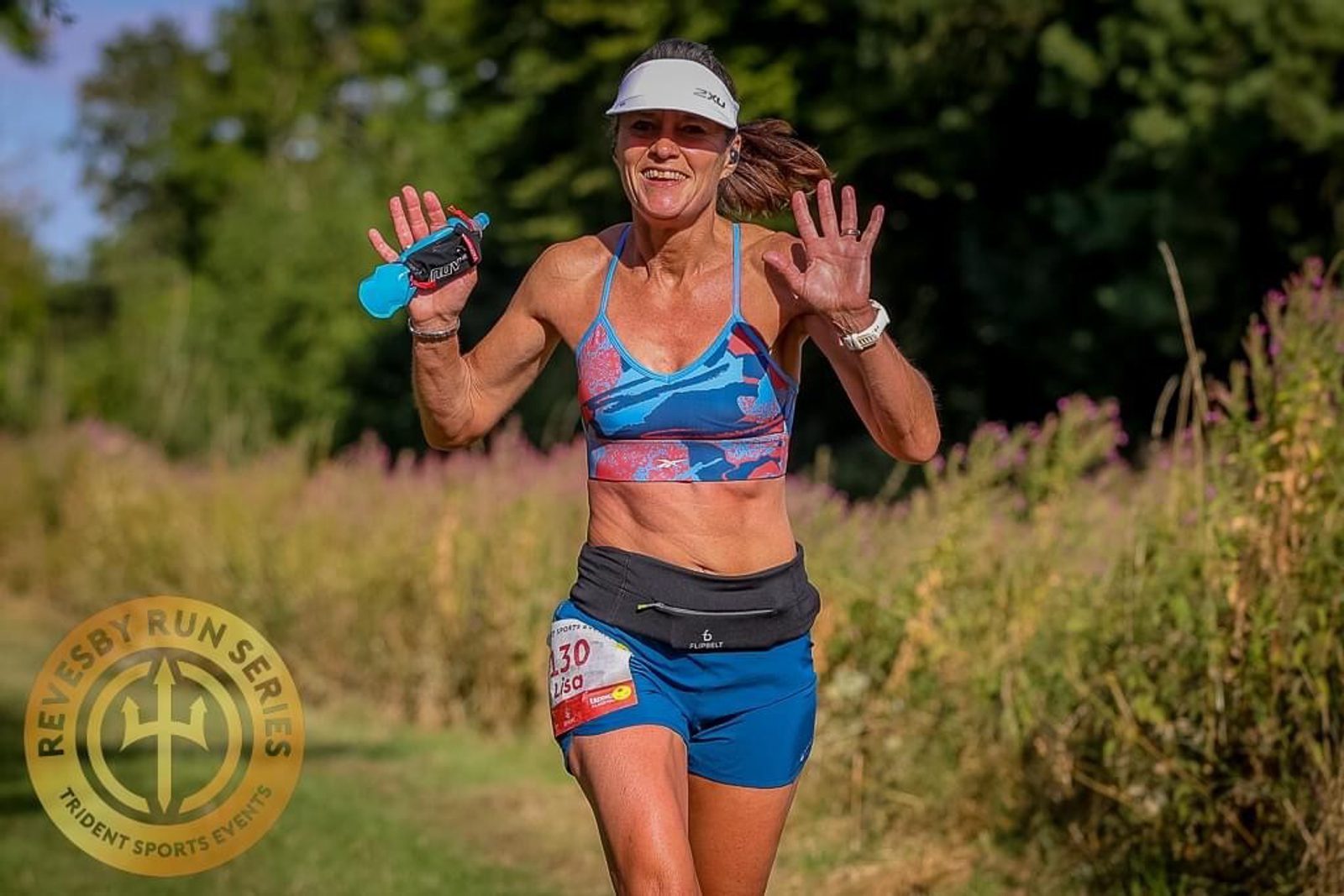 A woman in athletic attire and visor cap is smiling and raising her hands mid-run during an outdoor race. She is wearing a blue sports bra and shorts, with a race number attached to her waist. The background features lush greenery and the Revesby Run Series logo is visible.