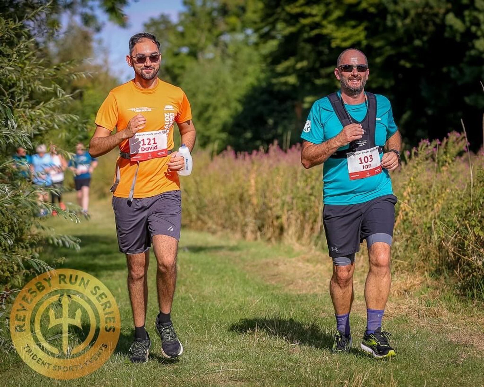 Two men are running outdoors on a sunny day. The man on the left in an orange shirt smiles and holds a water bottle, while the man on the right in a teal shirt and sunglasses also smiles. Both wear race bibs with numbers. Grass and trees are in the background.