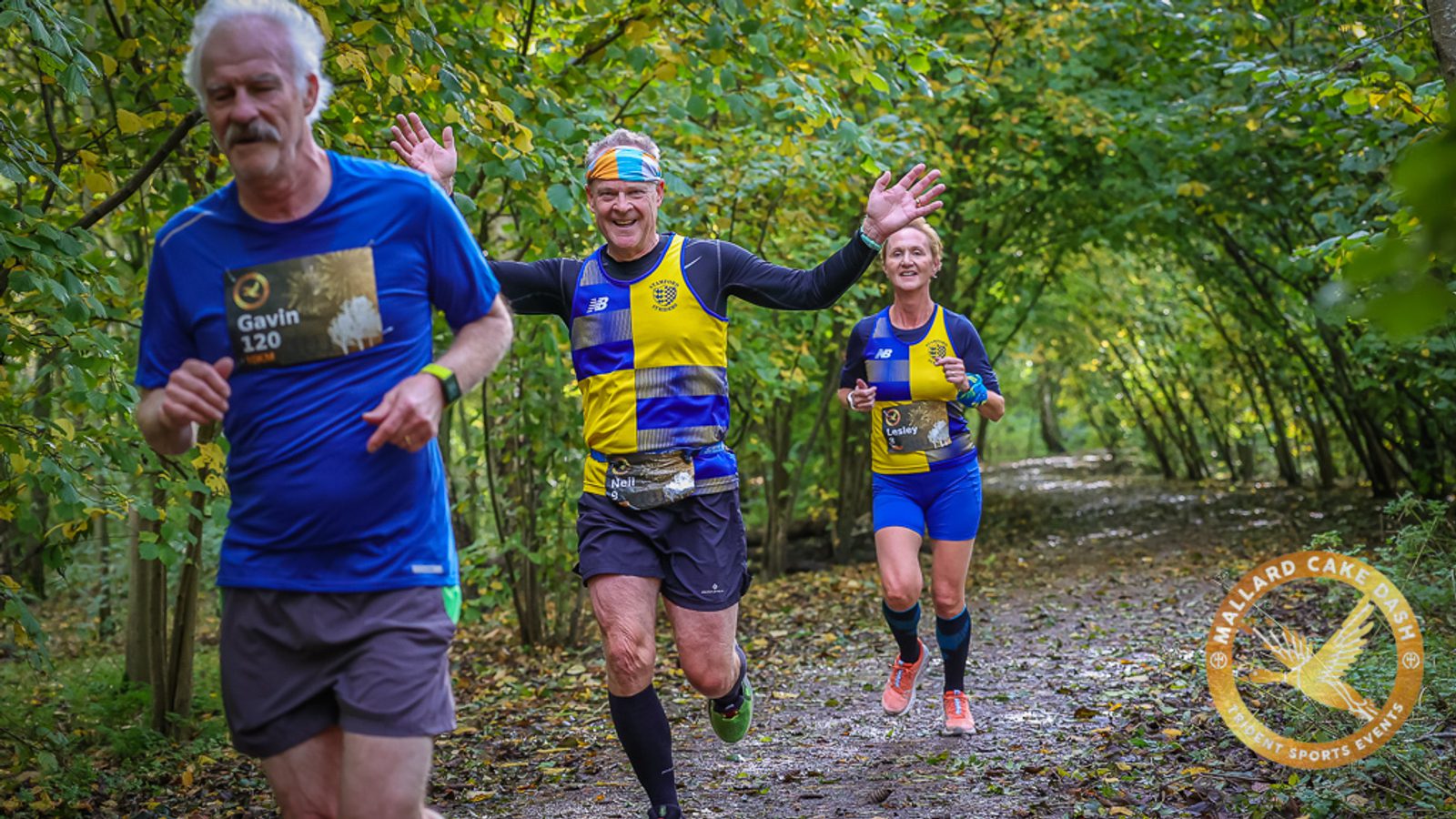 Three runners navigate a forest trail during a race. The runner in front, wearing a blue shirt labeled "Gavin 120," is followed by a man in a yellow and blue outfit excitedly raising his arms, and a woman, also in yellow and blue, smiling. Trees line the path.