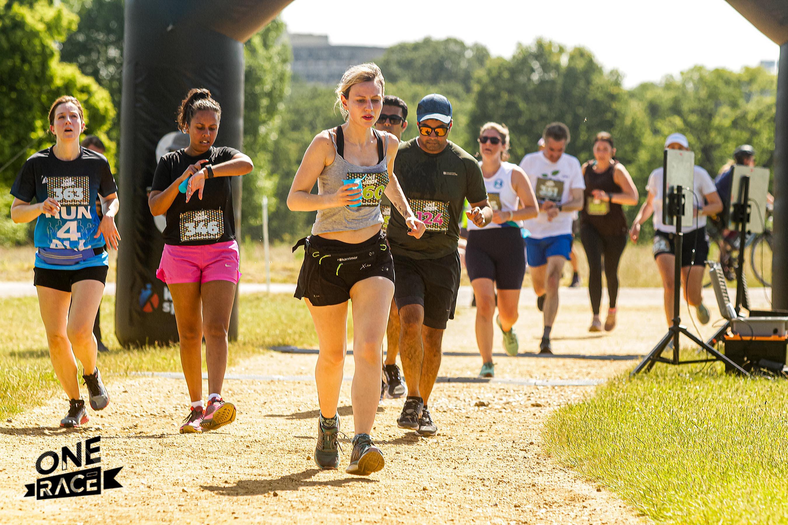 Runners of various genders and ethnicities participating in a marathon on a sunny day, with trees and a race arch in the background. Some wear bib numbers. A "One Race" logo is visible in the bottom left corner.