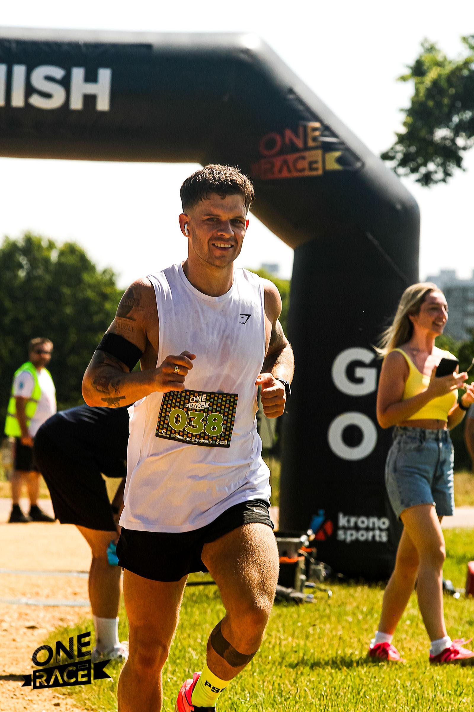 A runner in a white tank top and black shorts crosses the finish line at an outdoor race. A large black inflatable arch with "ONE RACE" and "GO" is in the background. A woman holds a clipboard, and the sun is shining brightly.