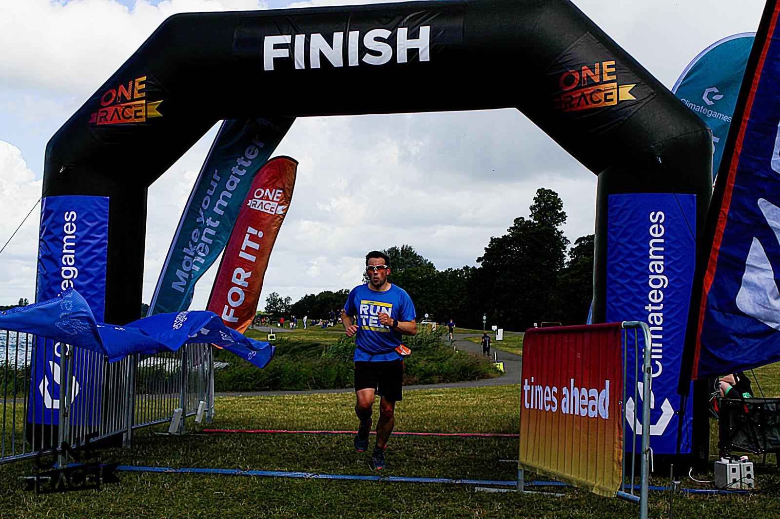 A runner in a blue shirt crosses a black finish line arch in an outdoor race. The arch has colorful sponsor logos. Flags and banners are displayed, and a few spectators are visible in the background under a partly cloudy sky.