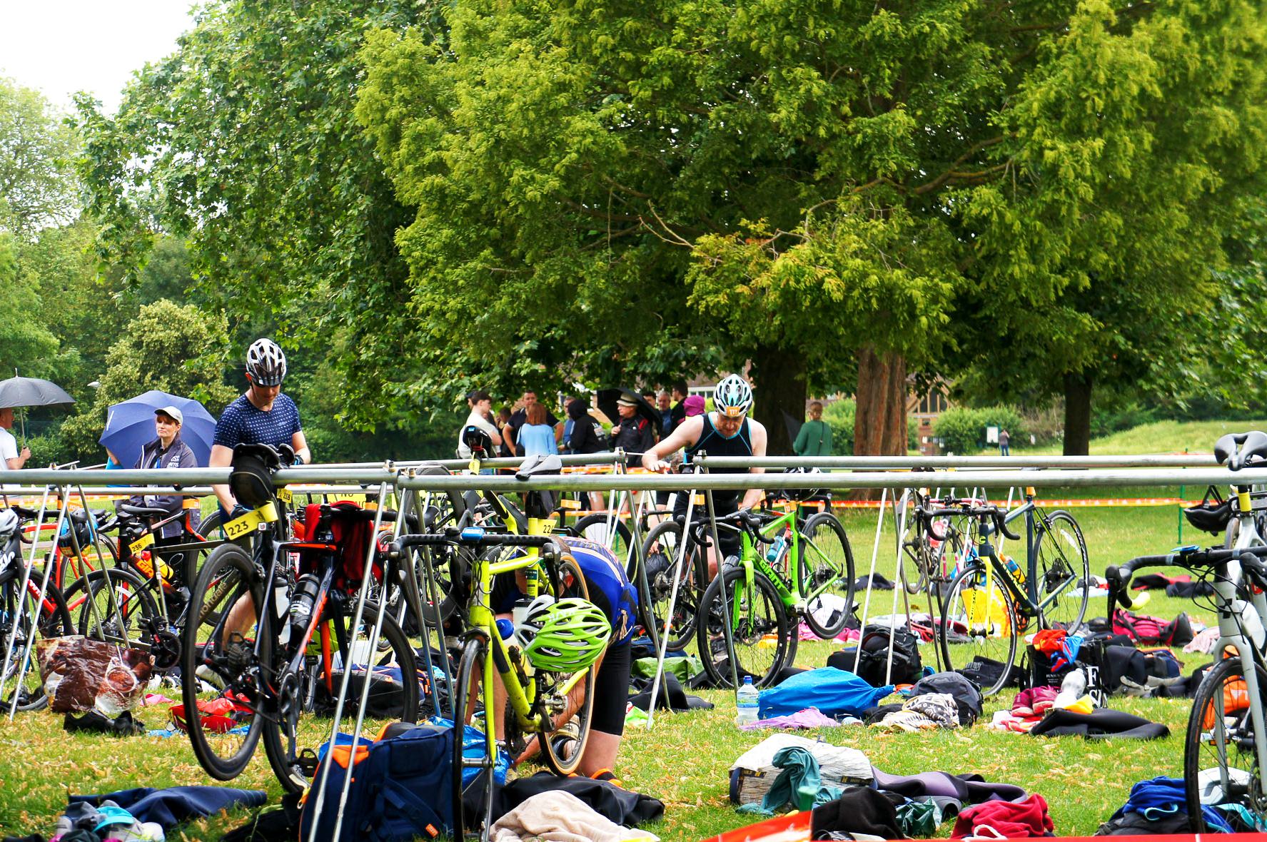 Cyclists transition area in an outdoor triathlon event. Participants wearing helmets prepare bikes and gear under trees. Various bicycles and equipment are scattered on the grass, with spectators in the background.