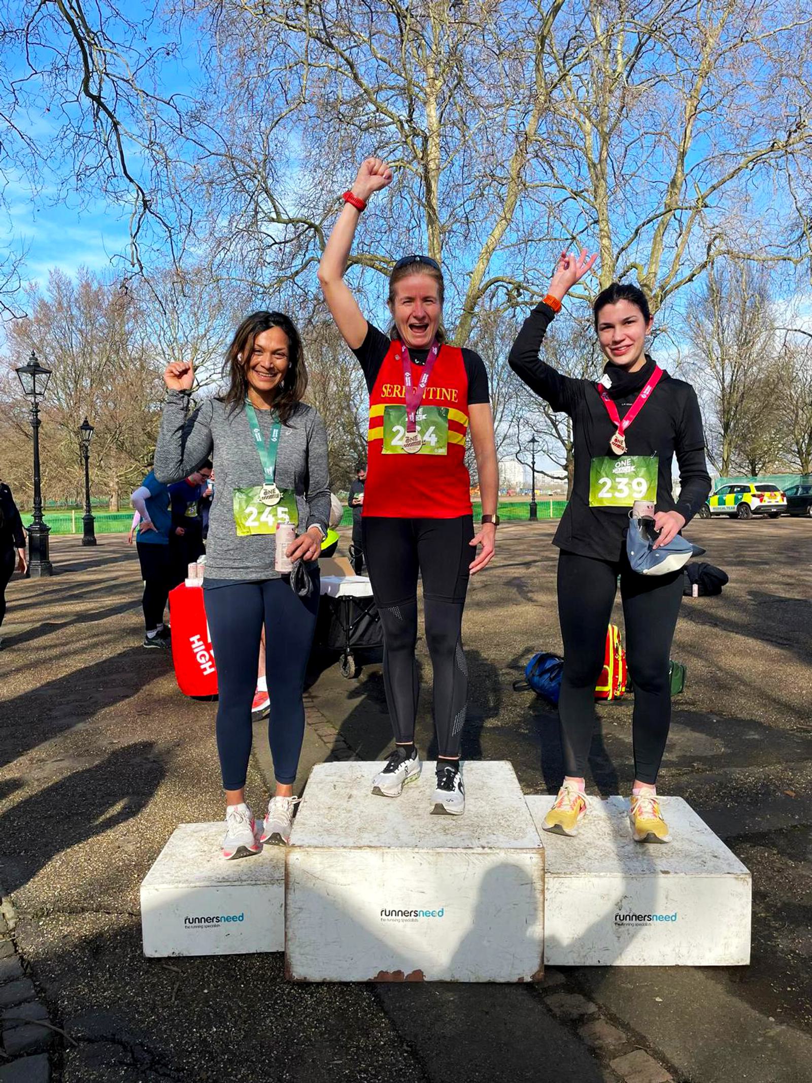 Three people stand on a winners' podium outdoors, each holding up a hand in celebration. The person in the center wears a red shirt, while the others wear black. They all have medals around their necks, and race numbers are visible on their shirts.