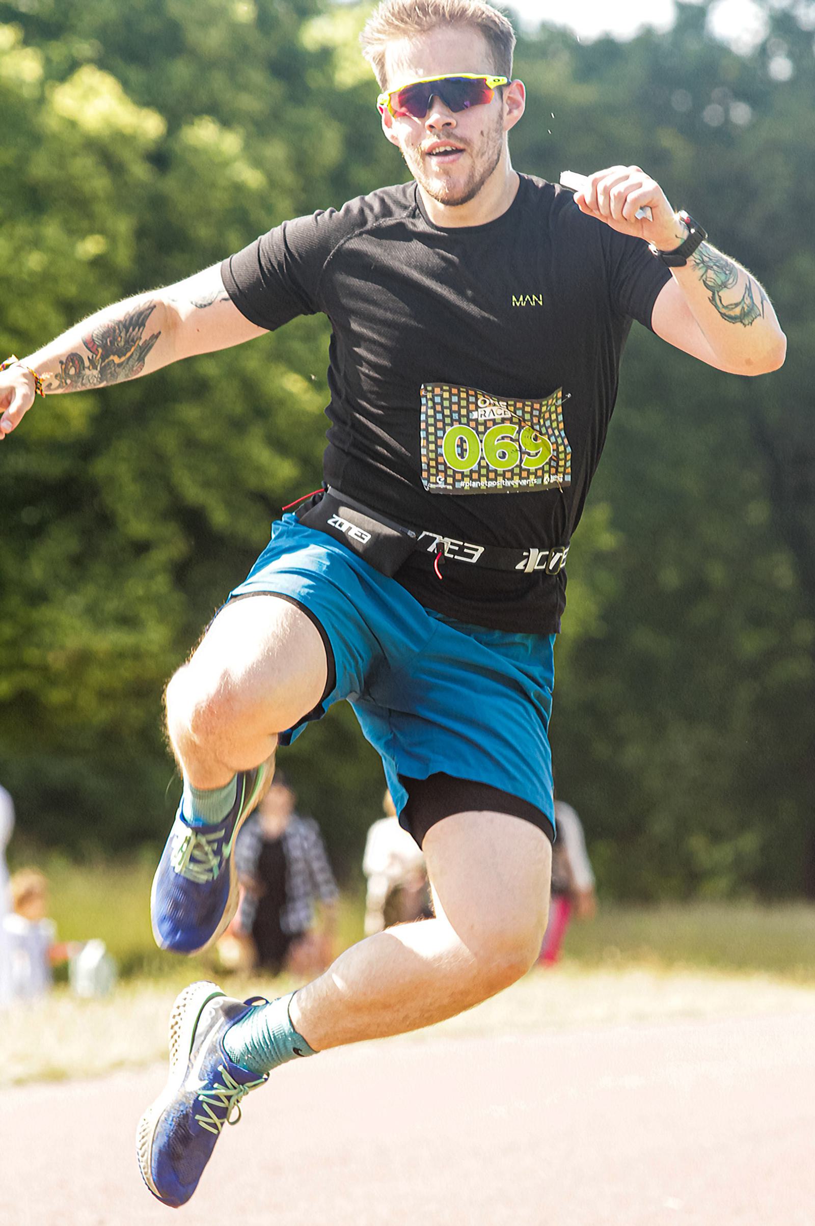 A man with tattoos, wearing sunglasses, a black shirt, blue shorts, and running shoes, is mid-jump on a track outdoors. He has a race number on his shirt and a belt around his waist. Trees are visible in the background.