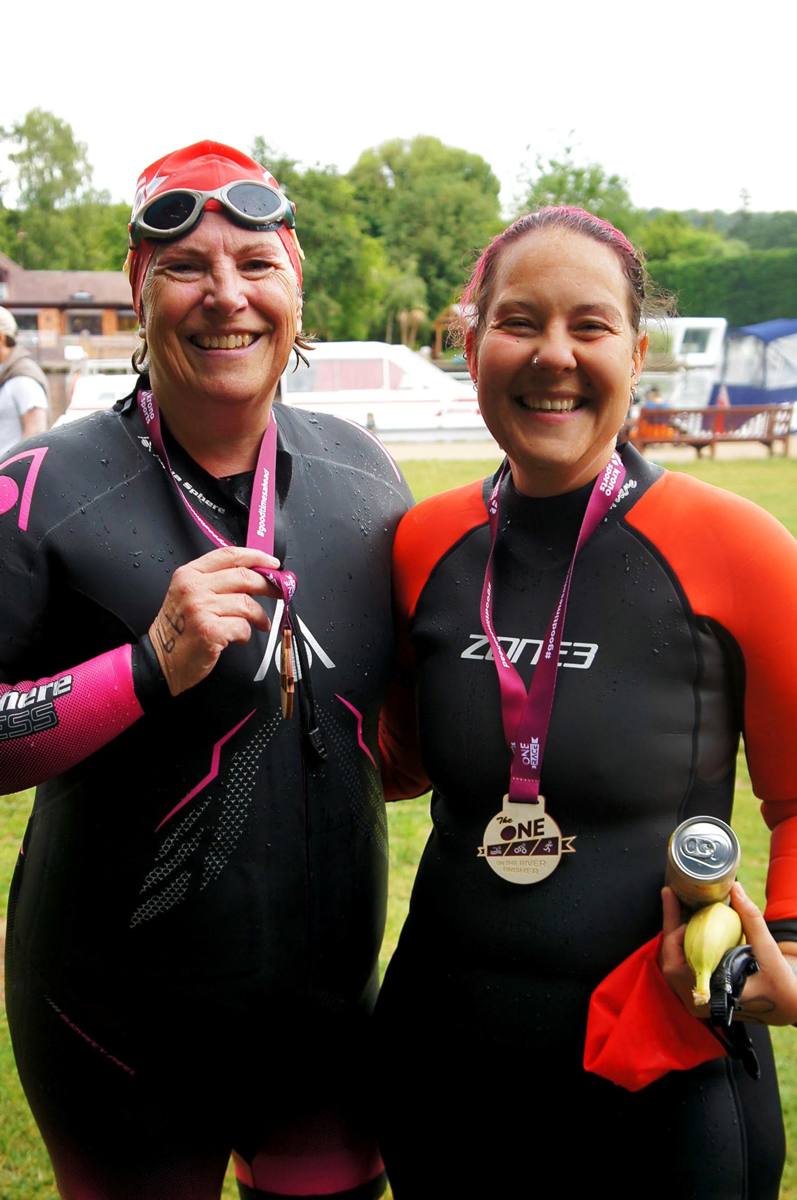 Two women in wetsuits and swim caps smiling, with medals around their necks, stand outdoors on grass. One holds a banana and water bottle. Trees and buildings are in the background.