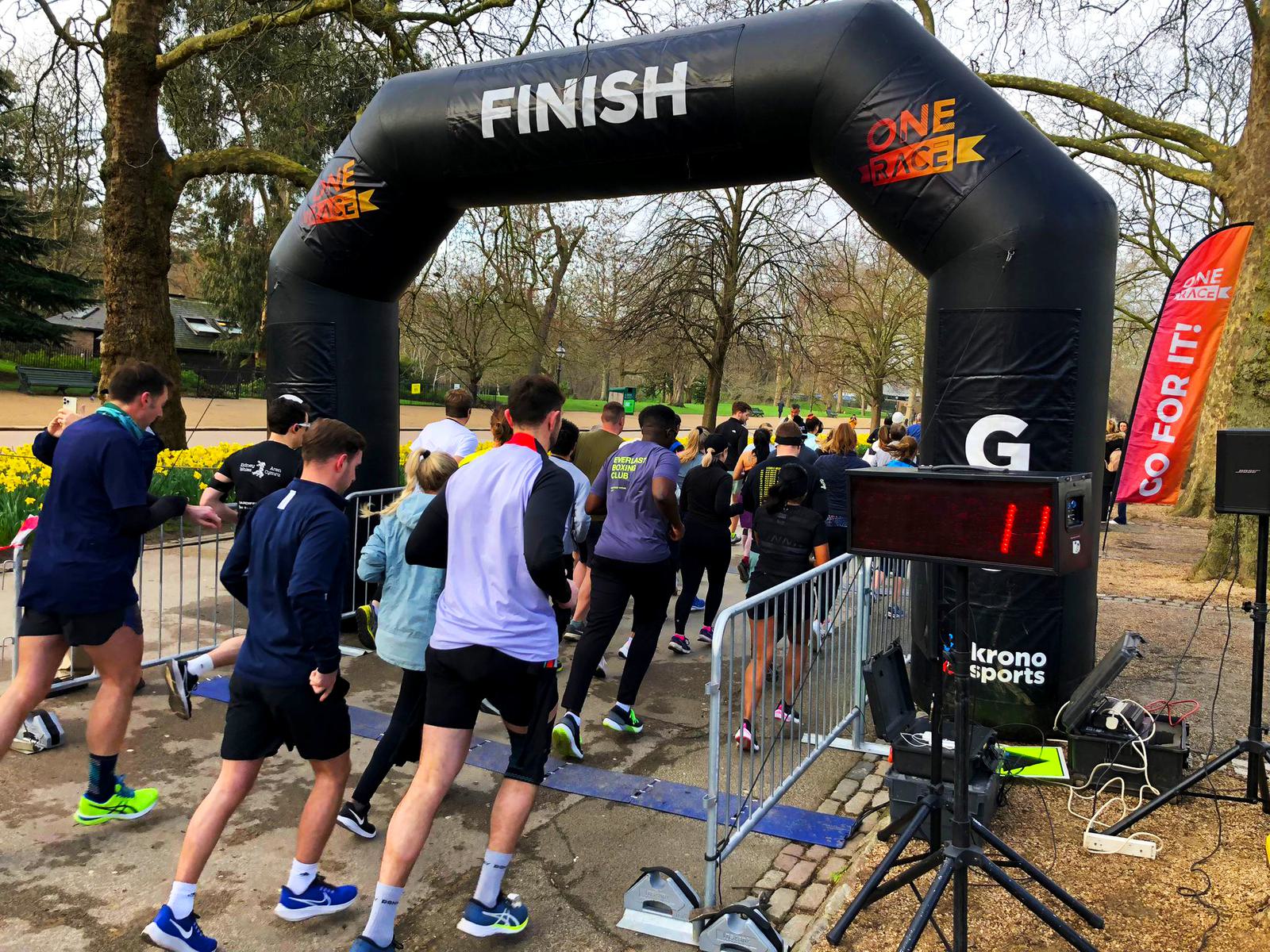 Runners cross the finish line at a race in a park. A black inflatable arch marked "FINISH" is in the background. A digital timer displays "11". Spectators watch from behind metal barriers. Trees and daffodils are visible in the background.