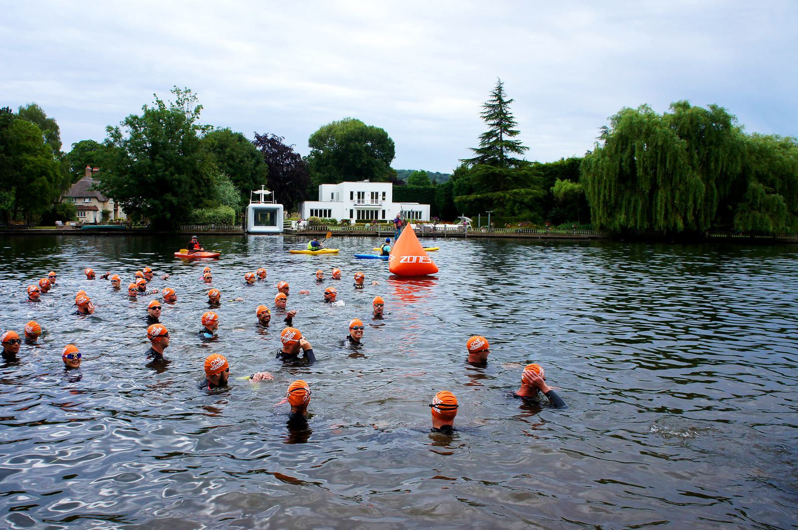 A group of swimmers wearing orange caps and wetsuits participate in an open water race. They are swimming in a river with a large, orange buoy marking the course. Trees and a white building are visible in the background. Kayaks are nearby.