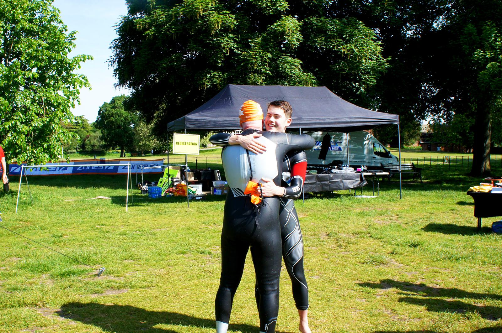Two people wearing wetsuits hug outdoors on a grassy field. A black canopy and a parked van are in the background, along with trees and a table with race signage. It appears to be a sunny day.