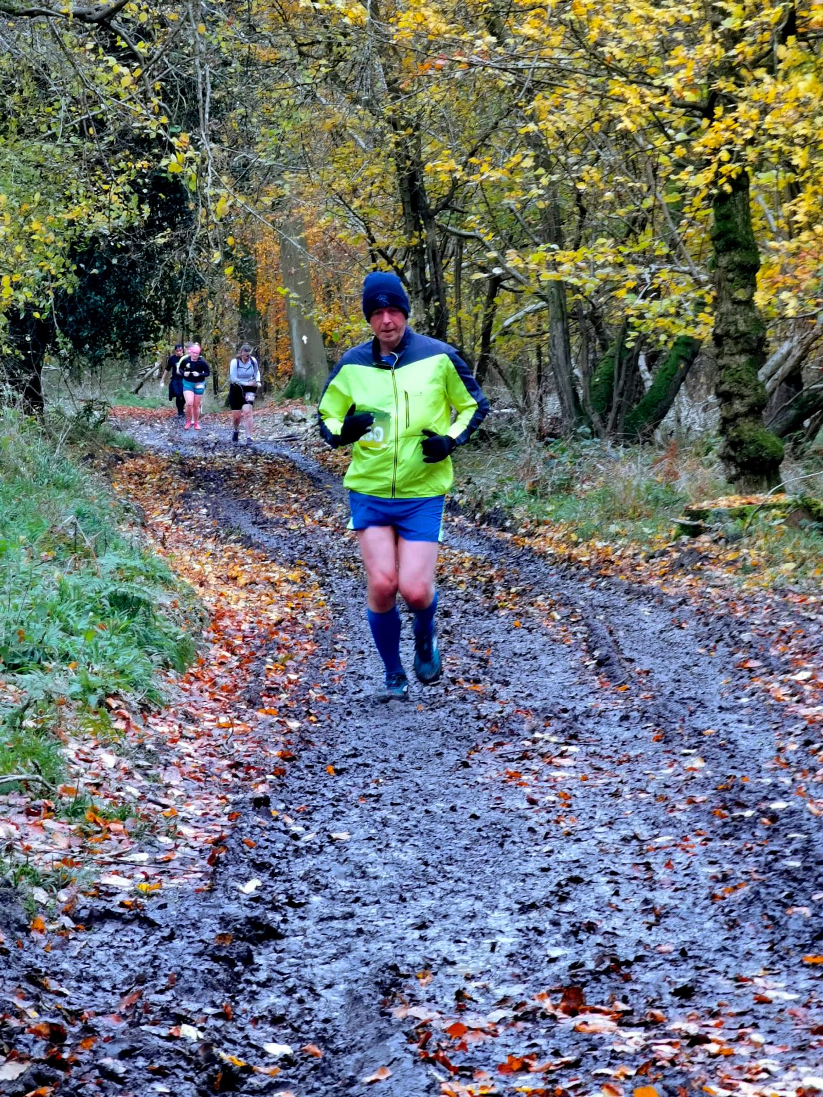 A person in a green and blue outfit jogs along a muddy forest trail surrounded by autumn trees. Three other people are running in the background, following the same path. The ground is covered with fallen leaves.