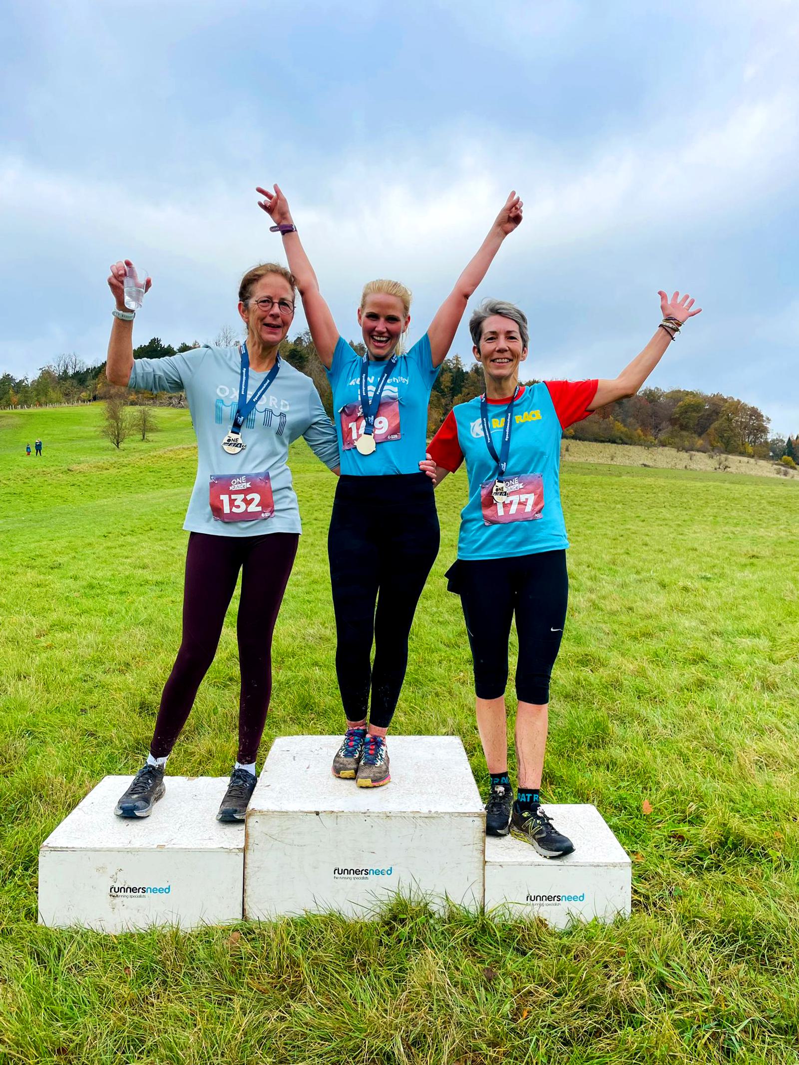 Three women stand on podiums in a grassy field, celebrating with arms raised. They're wearing medals and race bibs numbered 132, 119, and 177. The sky is cloudy and there are trees in the background.