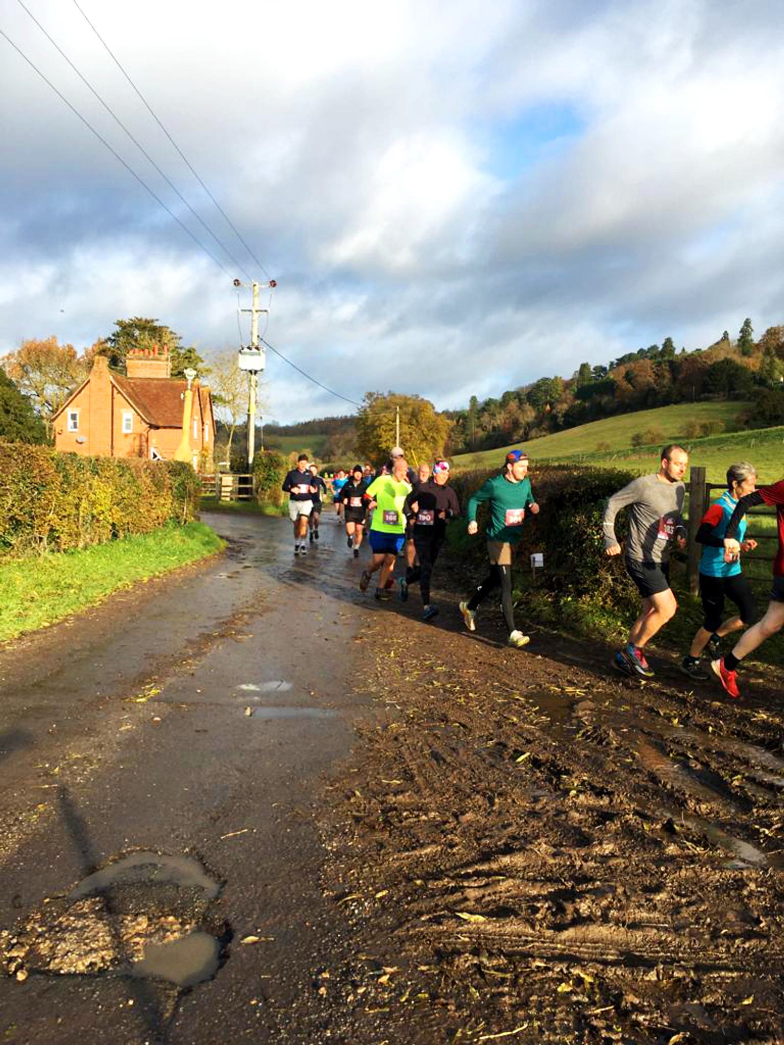 A group of runners on a countryside road during a race. The sky is partly cloudy, and the landscape includes a house, hedges, and fields. The road is wet with patches of mud. Participants wear various colorful running gear.