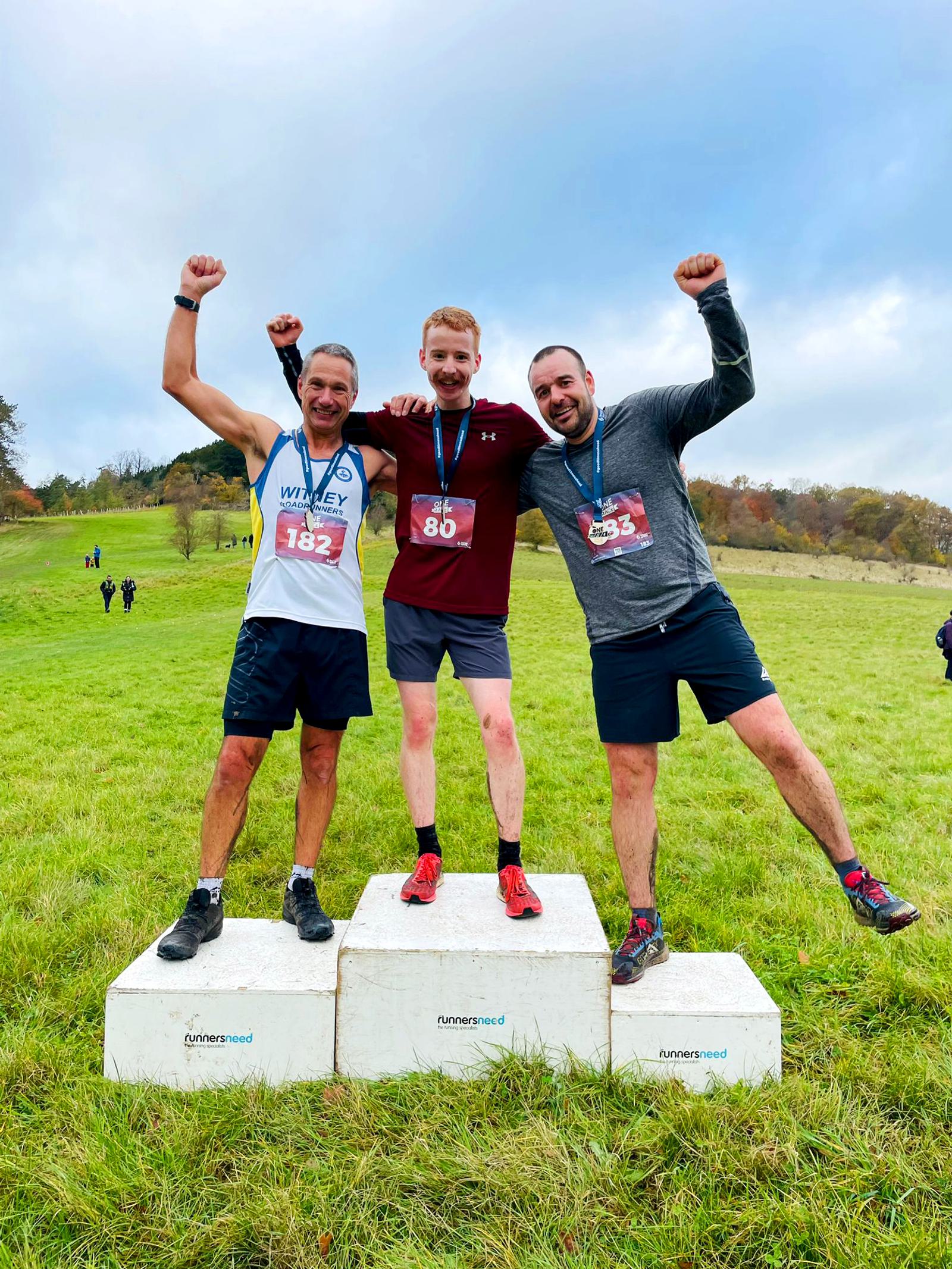Three men celebrate on podium blocks in a grassy field, wearing medals. The man on the left wears a white tank, the center man wears a red shirt, and the right man wears a gray hoodie. All have their arms raised joyfully.