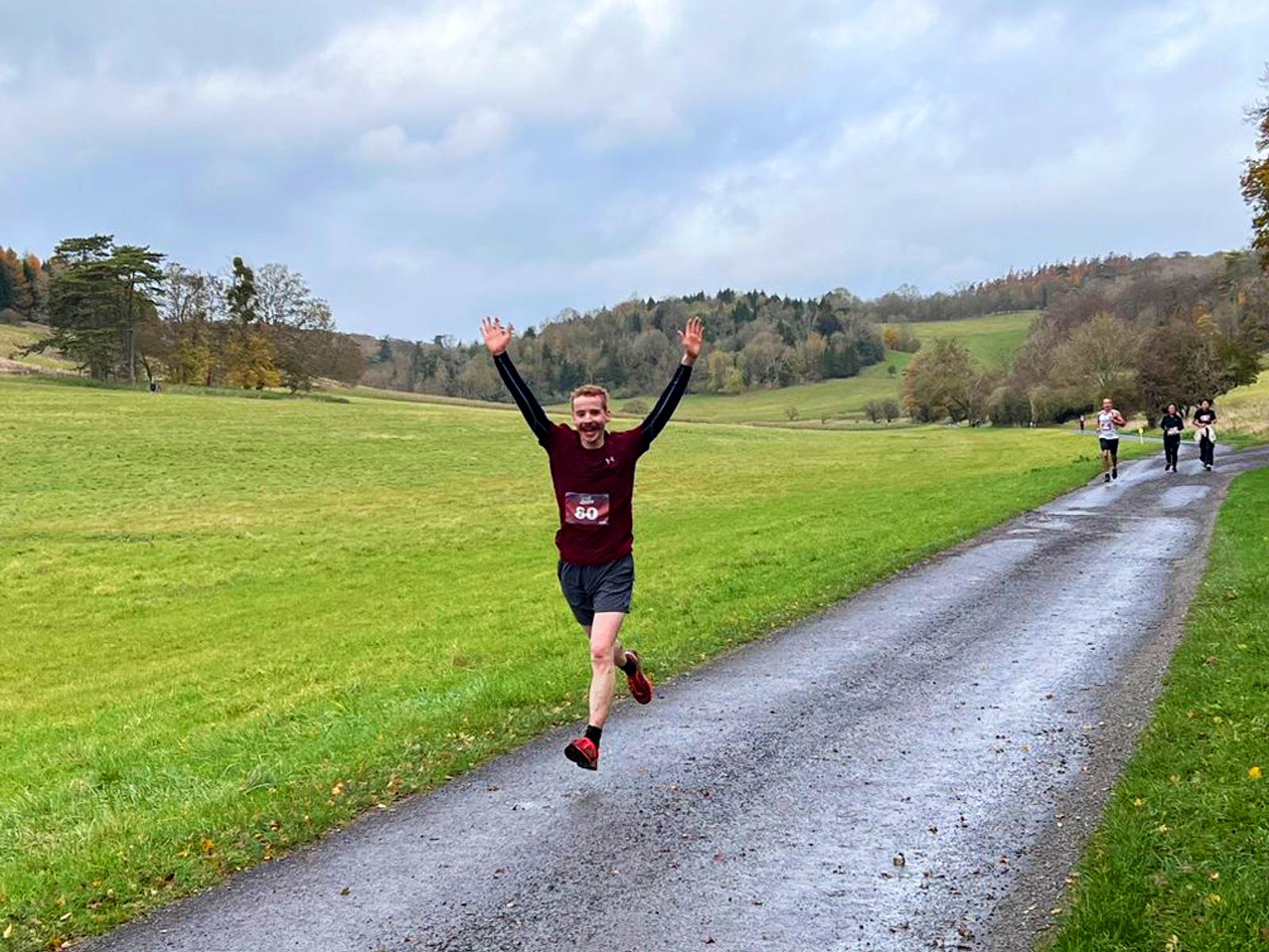 A person is running on a paved path in a park, smiling with arms raised. They're wearing a maroon shirt and shorts. The background features green grass, trees, and a cloudy sky. Other runners are visible in the distance.