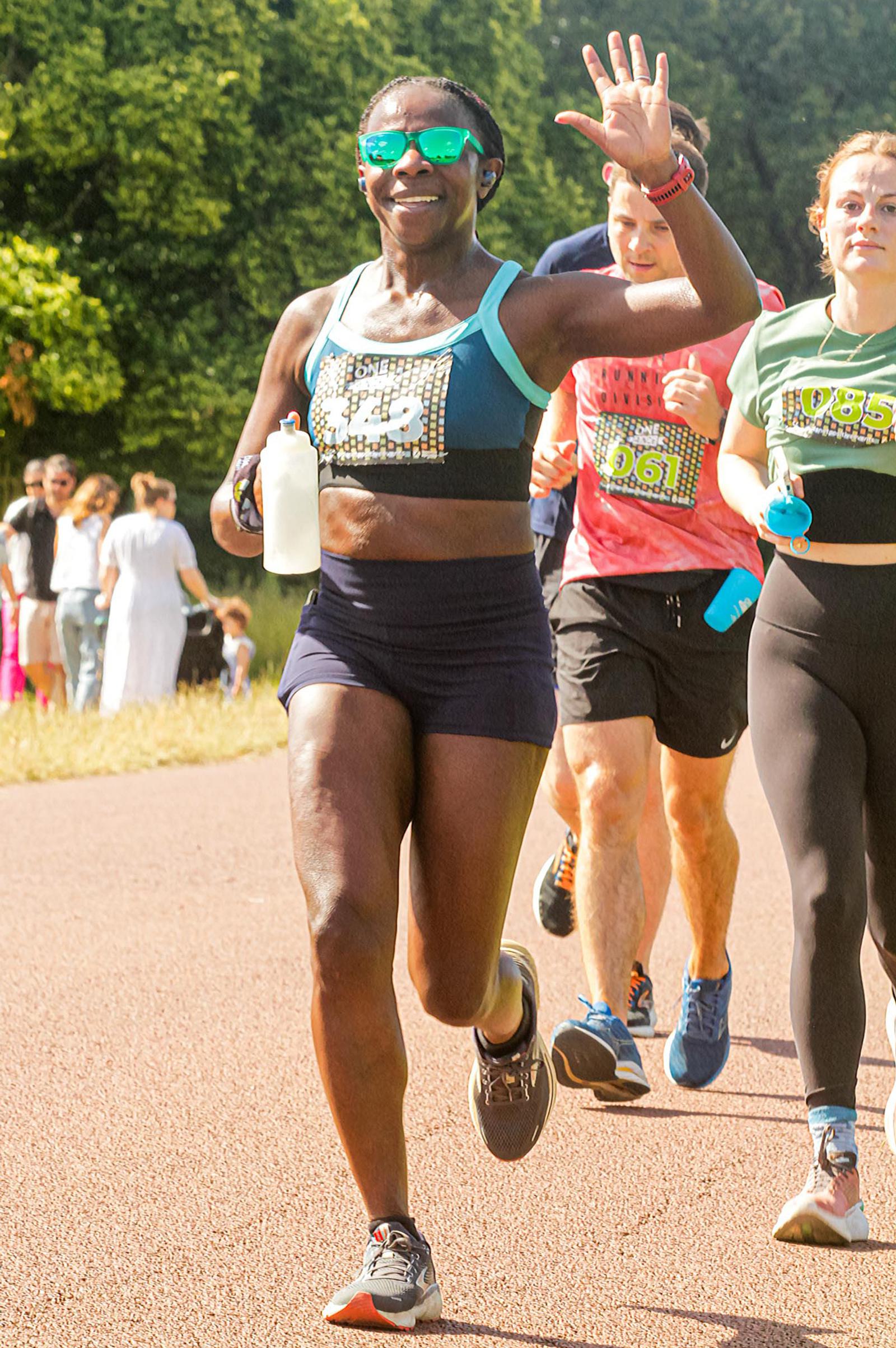 A woman in athletic wear and sunglasses waves cheerfully while running in a race, holding a water bottle. Other runners are beside her on a sunny day with people observing in the background.