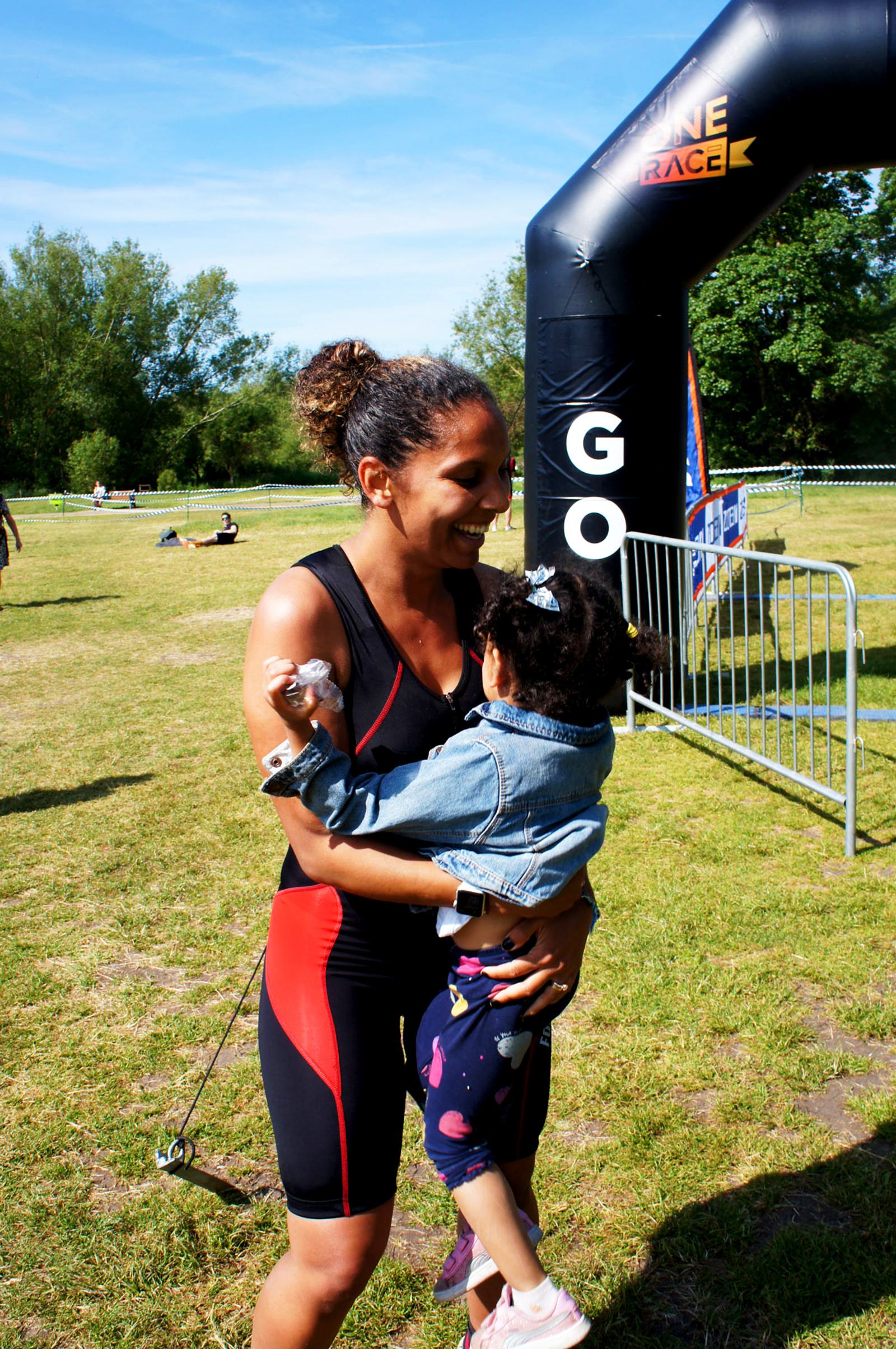 A woman in athletic gear joyfully holds a young child in her arms near the finish line of a race. They are outdoors on a grassy area with trees in the background, and a black inflatable arch marked "GO" is visible.