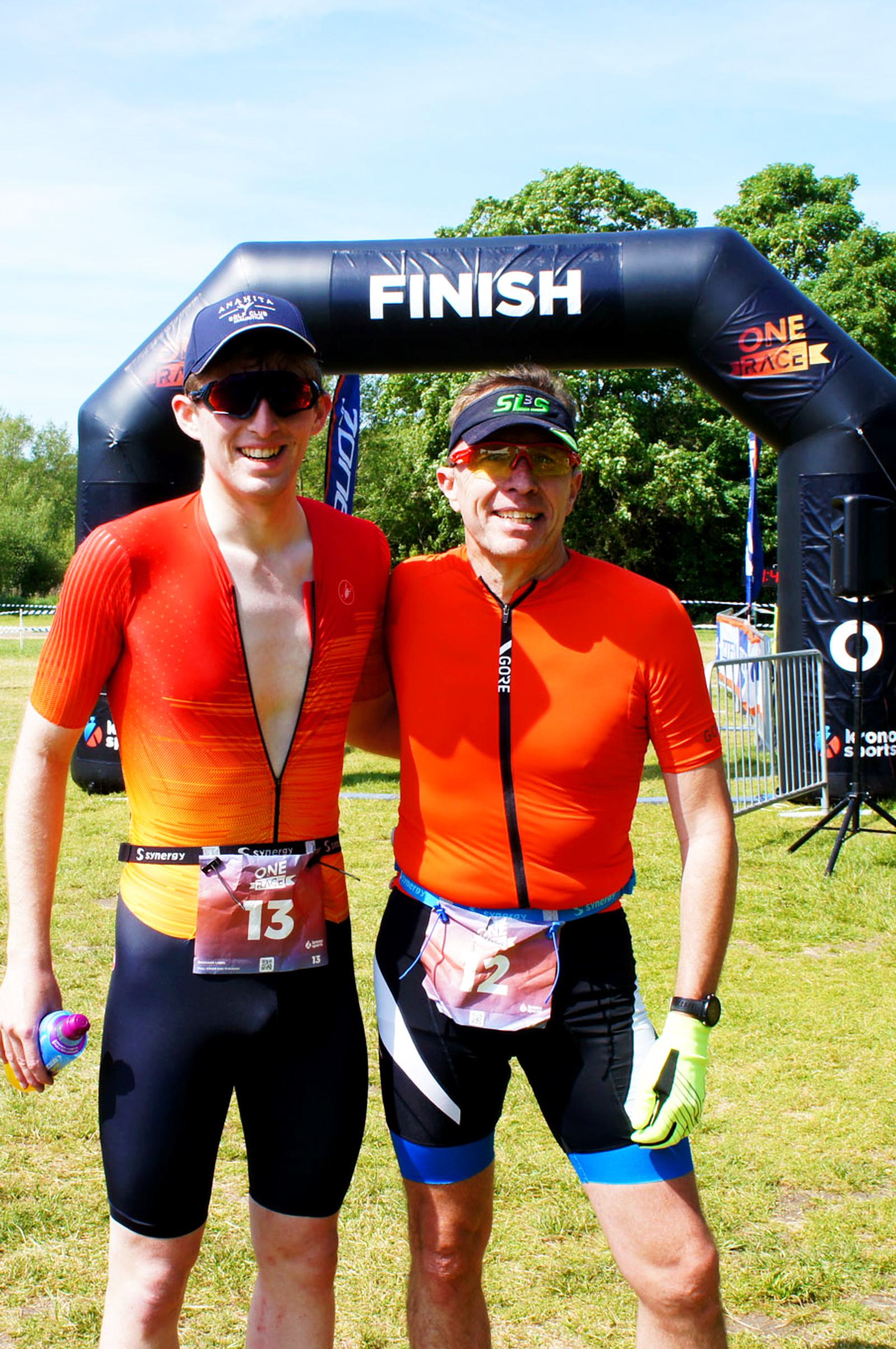 Two people in athletic gear stand smiling in front of a "Finish" arch at an outdoor race. One holds a water bottle. Both wear sunglasses and have race numbers on their shorts. It's a sunny day with green trees in the background.