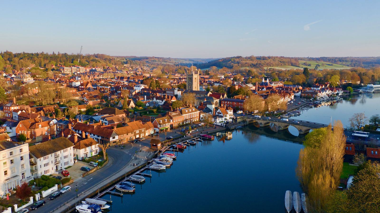 Aerial view of a picturesque town with red-roofed buildings along a river. Boats are docked at a marina, a stone bridge crosses the water, and lush greenery surrounds the area. A prominent church tower stands in the center, with hills in the background.