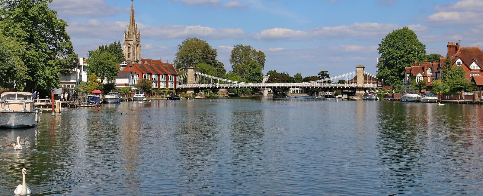 A scenic view of a riverside town featuring a suspension bridge, church steeple, and a mix of historic buildings. Boats are docked along the riverbank, and swans gracefully swim in the calm river under a clear blue sky with scattered clouds.