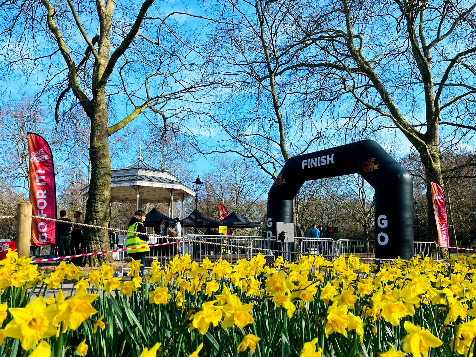 A park scene with a finish line archway and a gazebo. People are gathered near the structure, some in safety vests. Yellow daffodils are in the foreground, and bare trees stand under a clear blue sky.