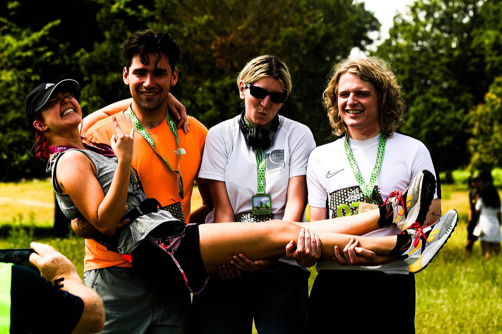 Four people in a park, smiling. Three are standing and holding up the fourth, who is lying horizontally and laughing. They wear casual athletic clothing and medals. Trees and greenery are in the background.