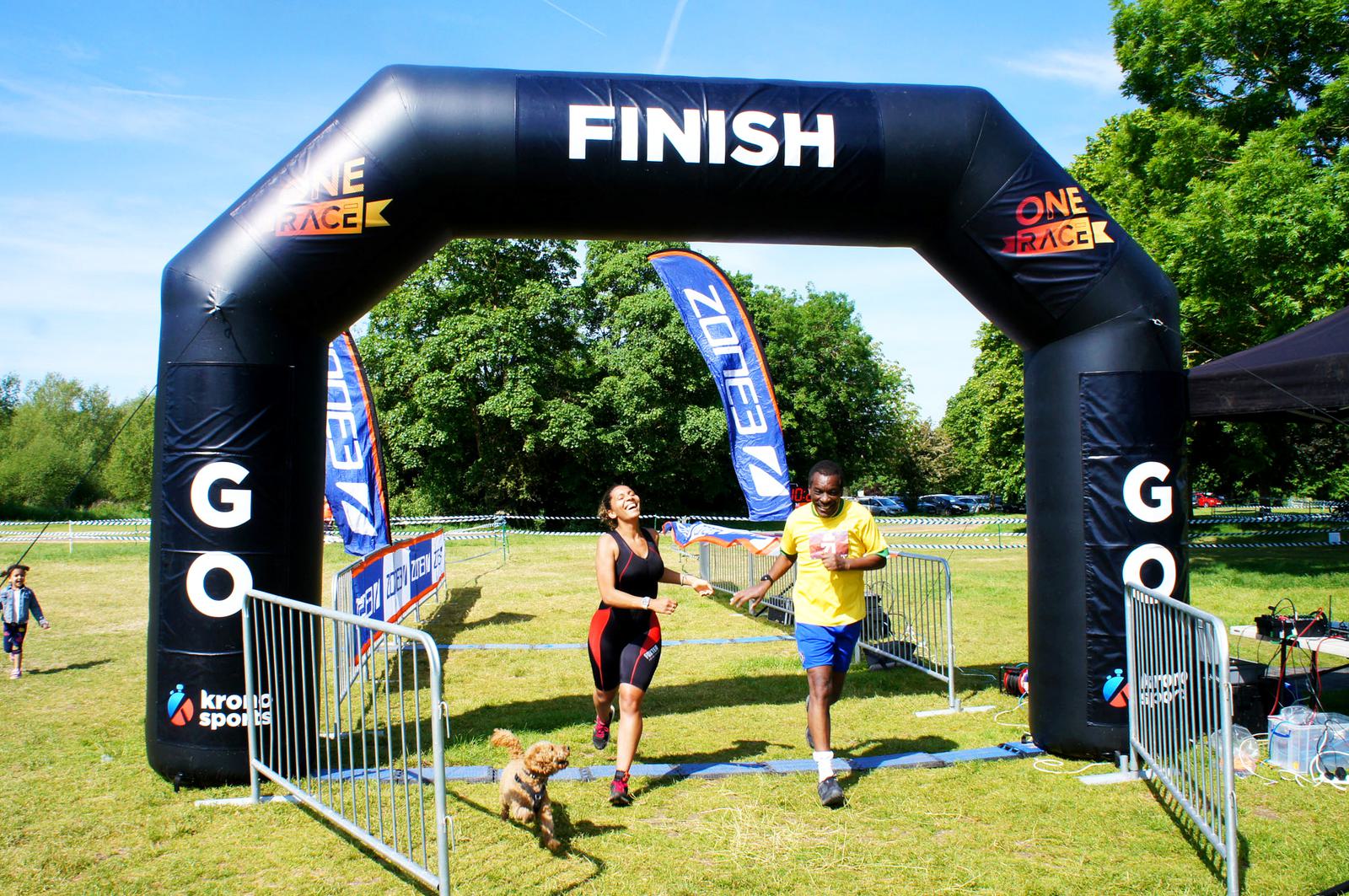 Two runners holding hands cross a finish line, smiling, under a black arch labeled "FINISH." A small dog runs beside them. Trees and banners are visible in the background, and a few people are nearby.