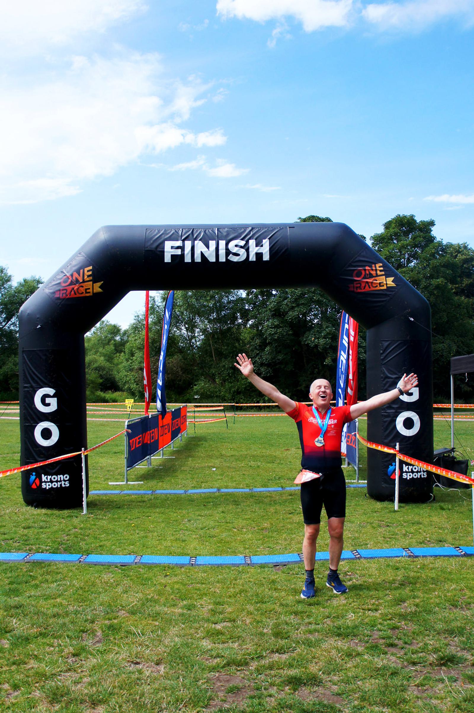 A person wearing a red shirt and shorts celebrates with arms raised under a black inflatable finish line arch at an outdoor racing event. The word "GO" is visible on the sides of the arch. Grass and trees are in the background.