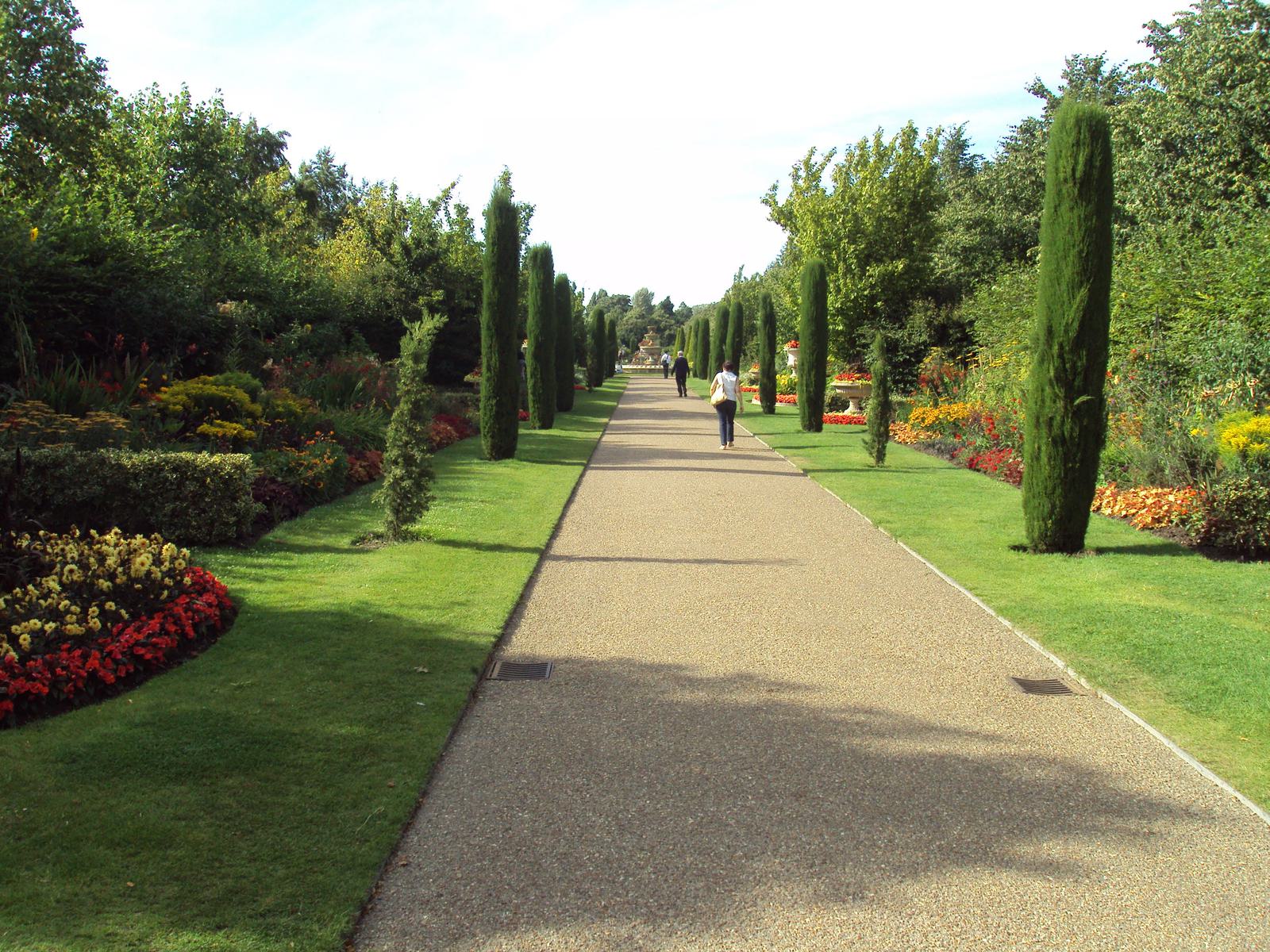 A long walkway in a garden with neatly trimmed grass on either side. Borders of colorful flowers and tall, slender trees line the path. A few people walk in the distance under a bright, clear sky.