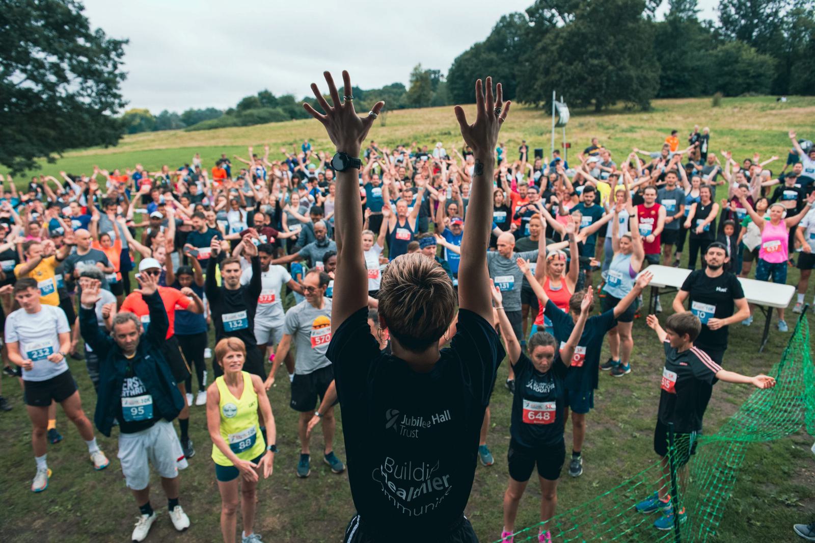 A large group of people participating in a pre-race warm-up outdoors is guided by an instructor on a raised platform. The participants, wearing numbered bibs, are engaged in stretching and raising their arms. The background shows a green park with trees and an overcast sky.