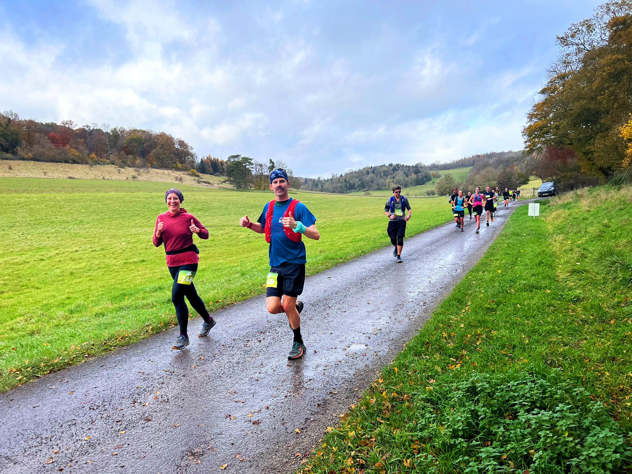 Runners on a country road participate in a race. The road is wet, suggesting recent rain, and is bordered by green fields. Trees with autumn foliage line the background, and the sky is partly cloudy. The participants wear athletic gear and race bibs.
