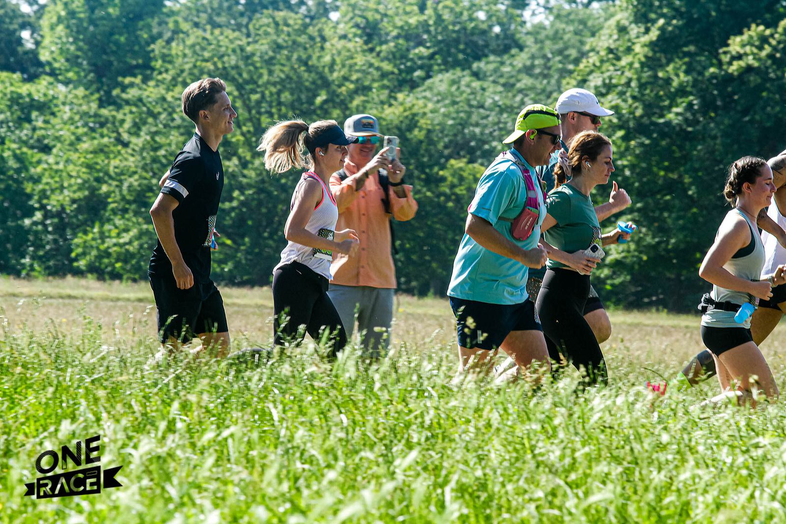 A group of people running through a grassy field with trees in the background. Some are wearing athletic gear, and one person is taking photos. The logo "ONE RACE" is in the bottom left corner.