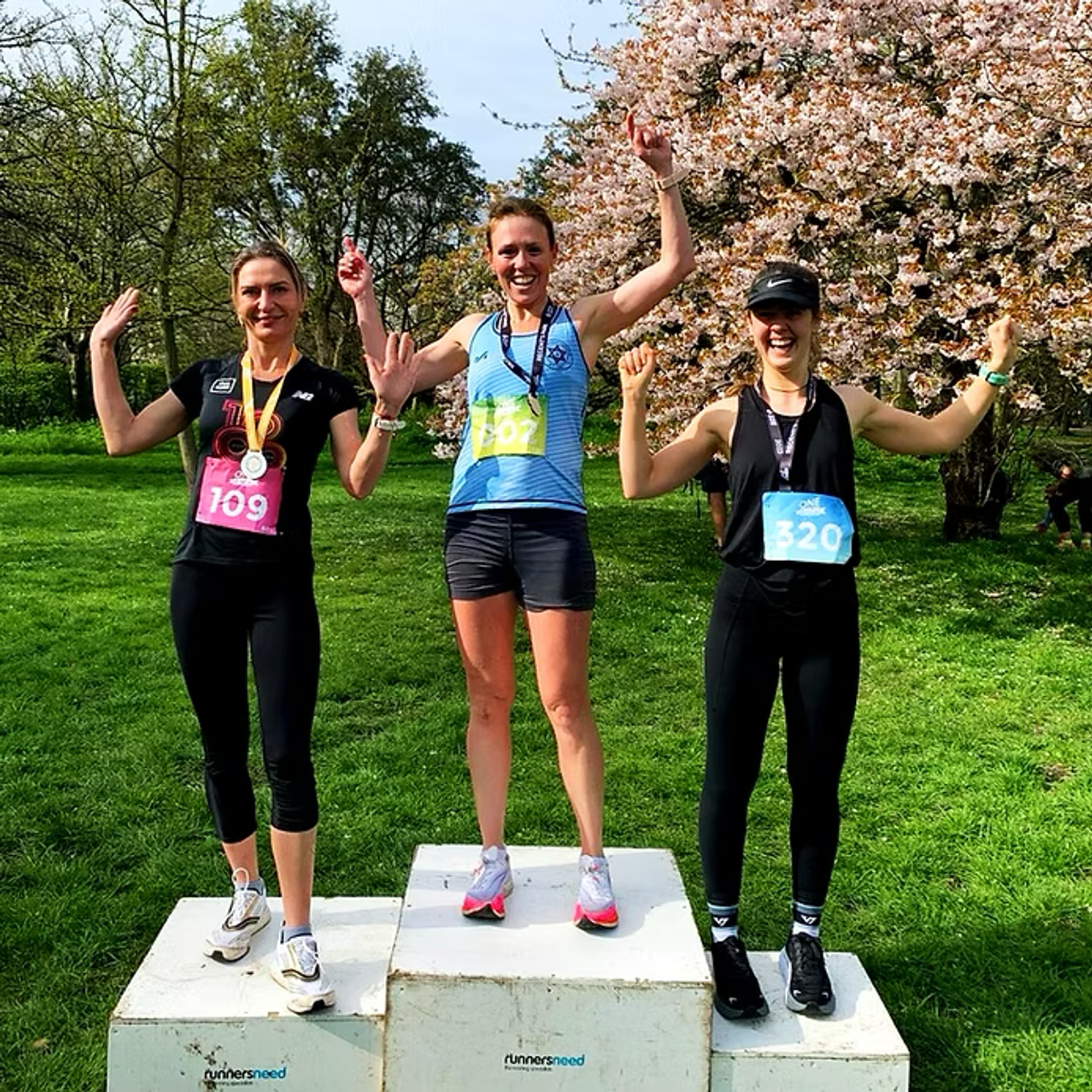 Three women celebrating on a winners' podium outdoors under a blooming tree. Each wears a medal and a race bib with numbers 109, 107, and 320. They smile and hold their arms up in victory, surrounded by greenery.
