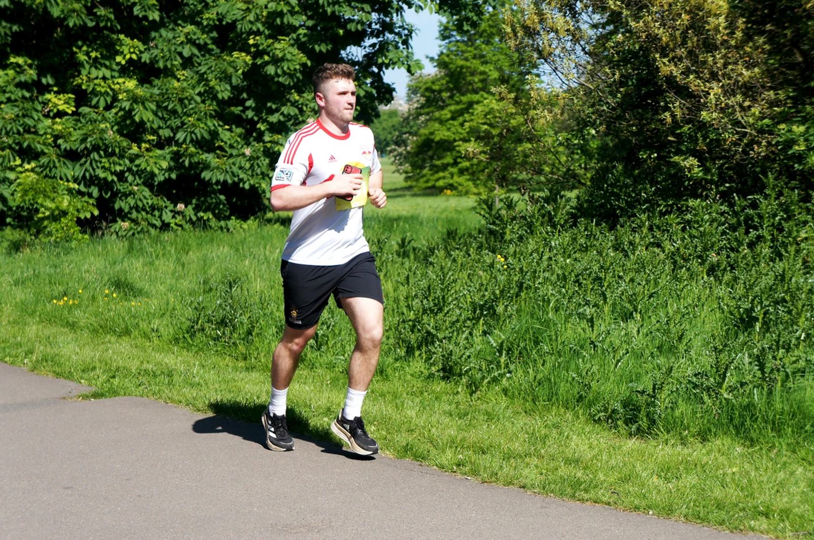 A person is jogging on a paved path in a park. They are wearing a white and red sports jersey, black shorts, and running shoes. The park is lush with greenery and trees under a clear blue sky.