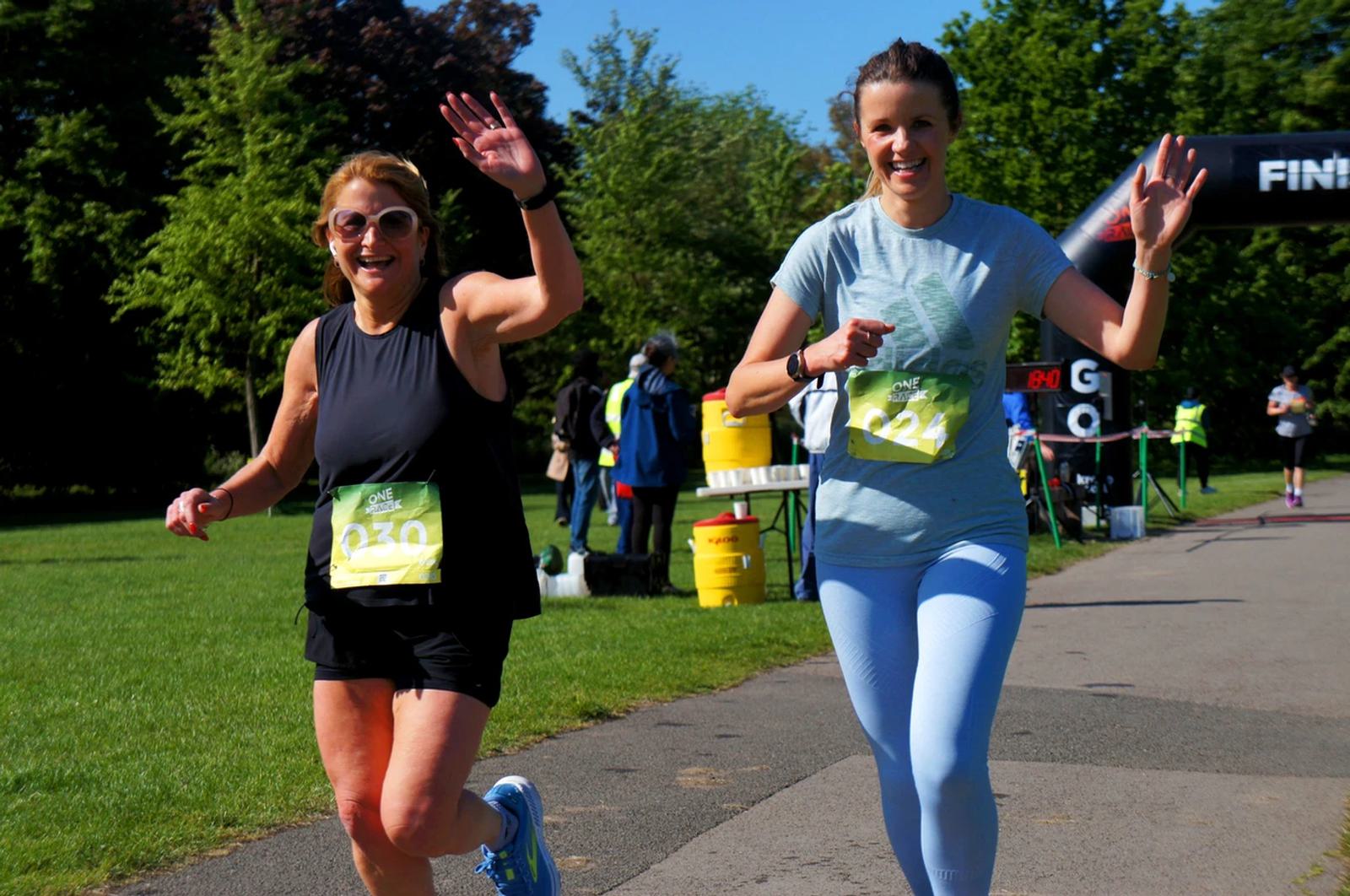 Two women jogging and waving cheerfully, wearing race bibs 230 and 224. The scene is outdoors on a sunny day, with green trees in the background and a finish line ahead. The atmosphere is lively and energetic.