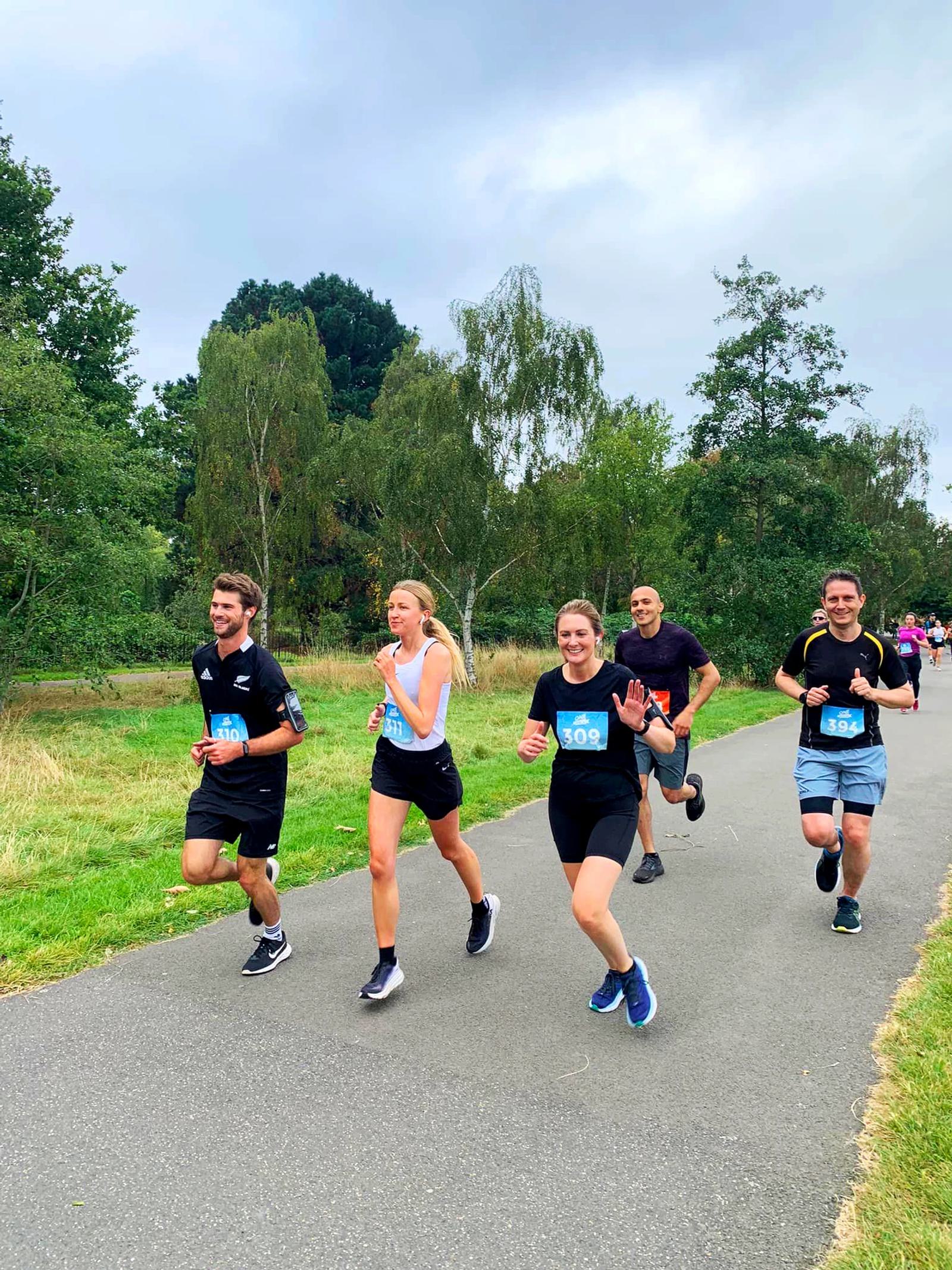 A group of six people running on a paved path in a park. They are wearing athletic clothing and numbered bibs. Trees and grass surround the area under a cloudy sky. Everyone appears to be smiling and enjoying the run.