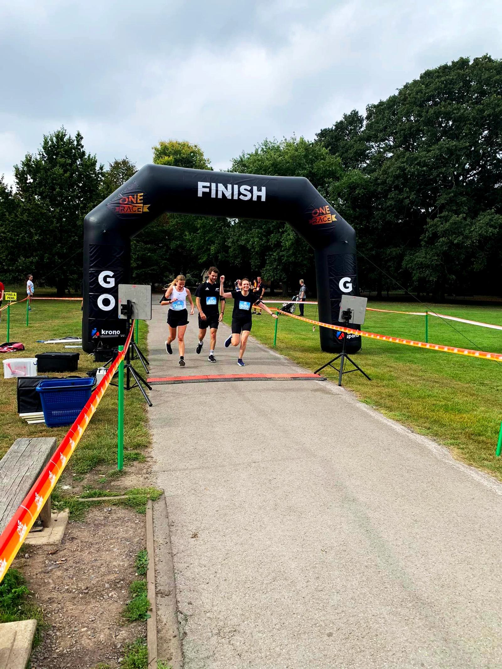Three people crossing a finish line together, celebrating with raised arms. They are on a paved path outdoors, surrounded by green grass and trees. An inflatable arch with the word "FINISH" marks the end of the race.