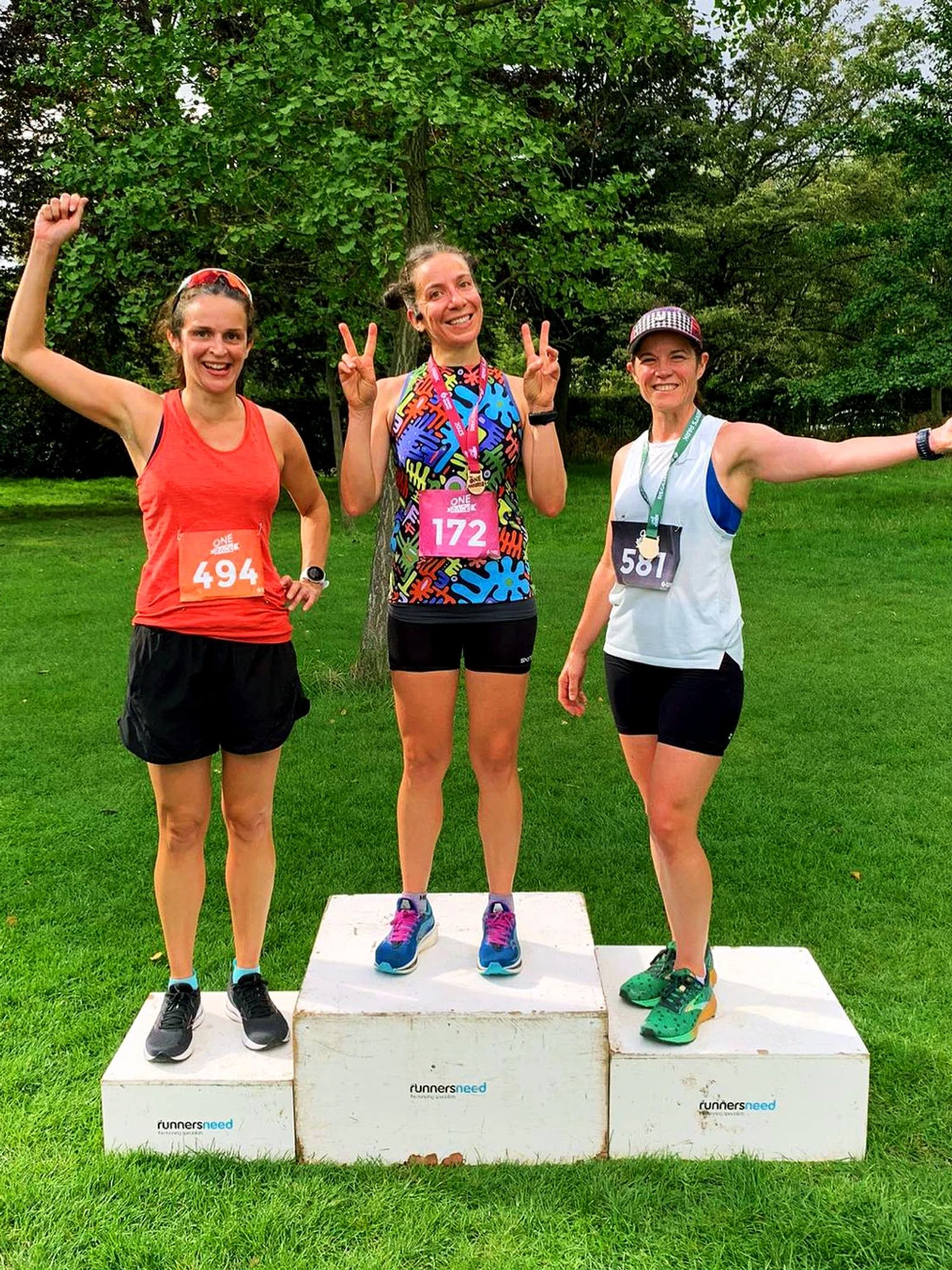 Three women stand on a winners' podium outdoors, smiling and celebrating. The woman on the left wears a red tank top, the center woman wears a colorful top, and the woman on the right wears a white tank top. Trees and grass are in the background.