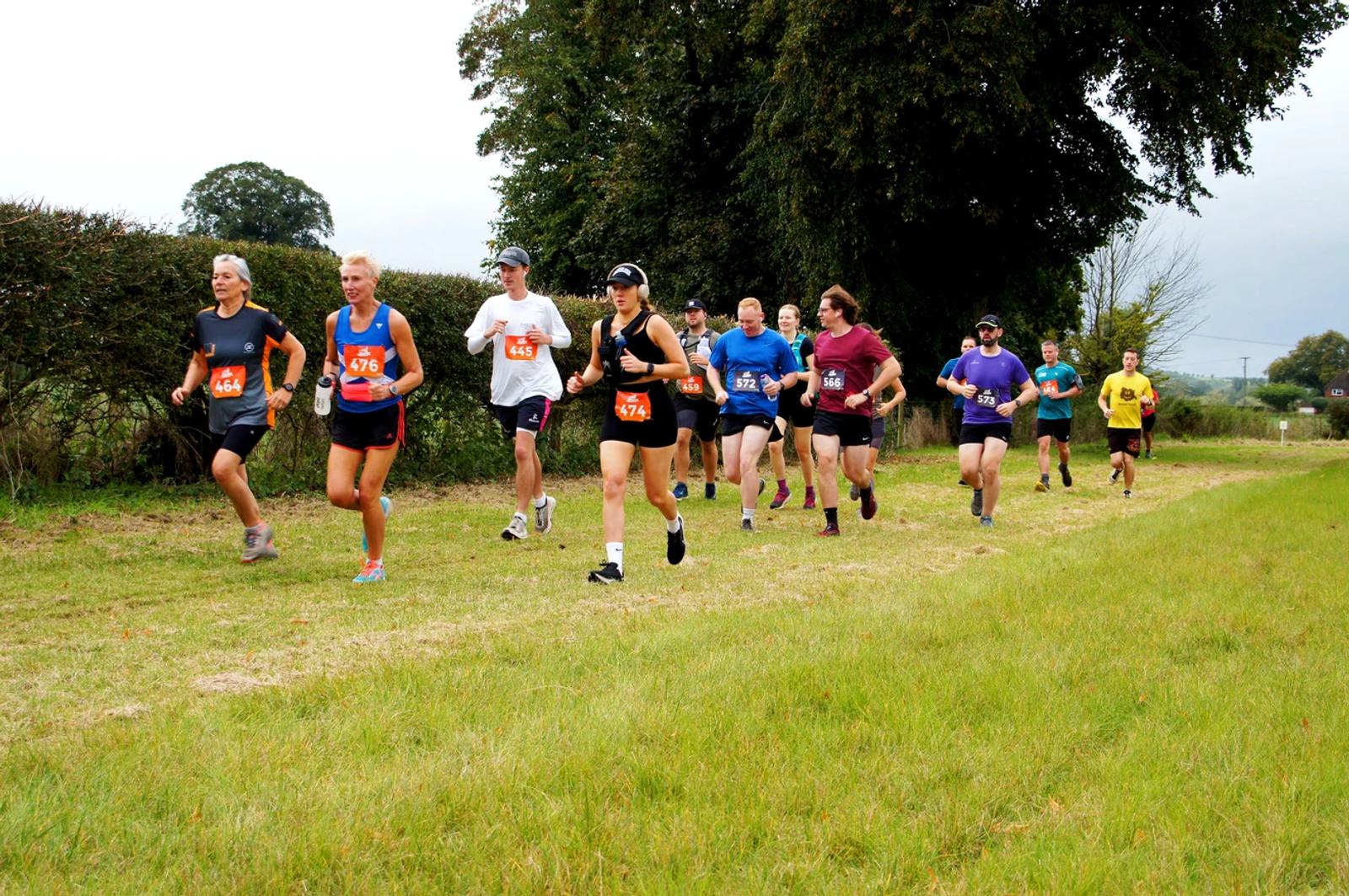 A group of runners is participating in an outdoor race, moving along a grassy path bordered by trees and hedges. They are wearing numbered bibs and athletic attire. The sky is overcast.