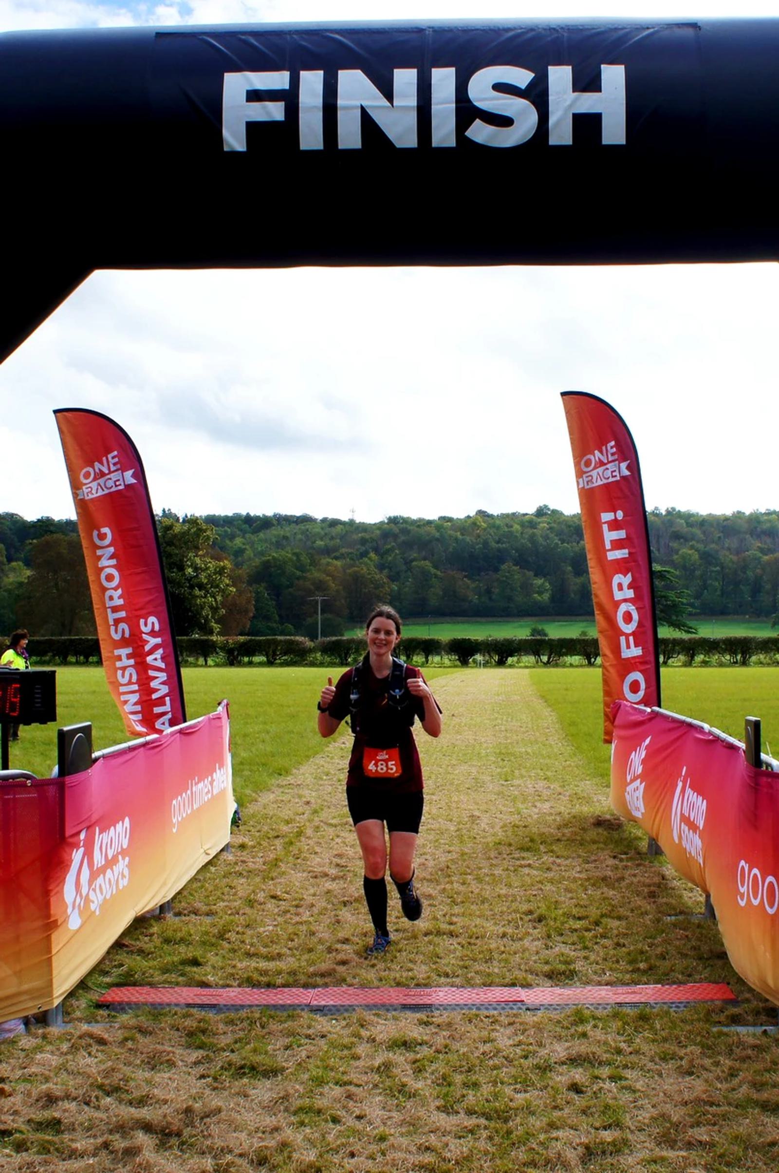 A runner wearing a bib crosses the finish line of a race. The finish banner stretches overhead with motivational flags on both sides. The background shows a grassy field and trees under a cloudy sky.