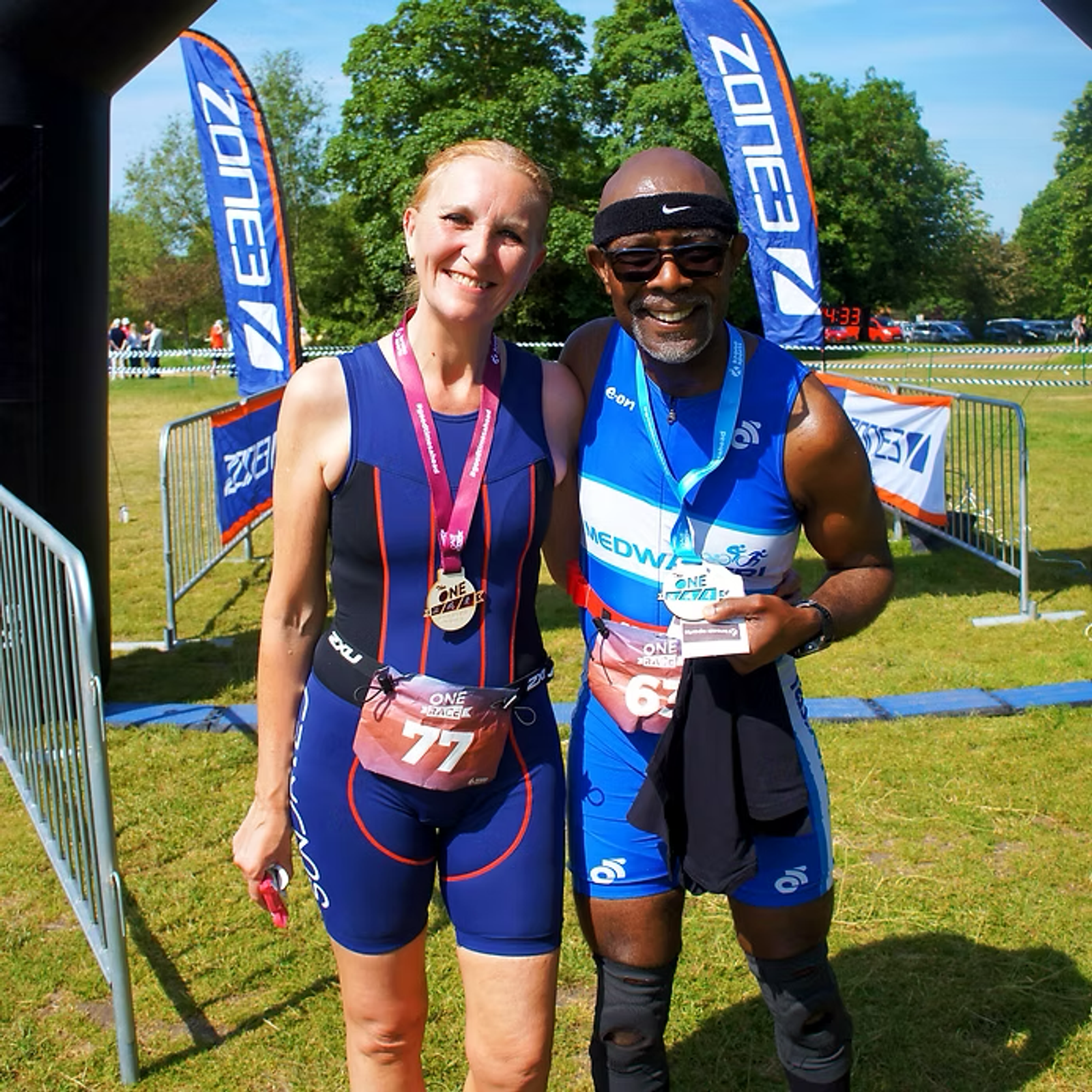 Two athletes wearing medals and race bibs are smiling and standing together at a finish line. The man on the right holds a trophy. They are outdoors on a sunny day with banners and green trees in the background.