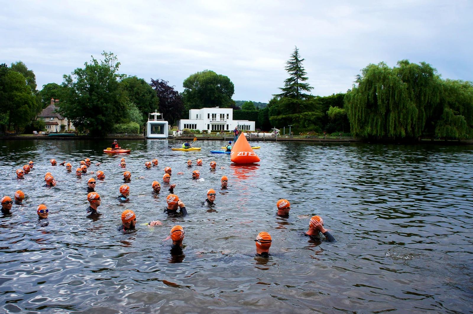 A group of swimmers wearing orange caps participate in an open water swim near a large orange buoy. Kayakers in the background oversee the event. The setting is a river with lush greenery and houses on the banks.