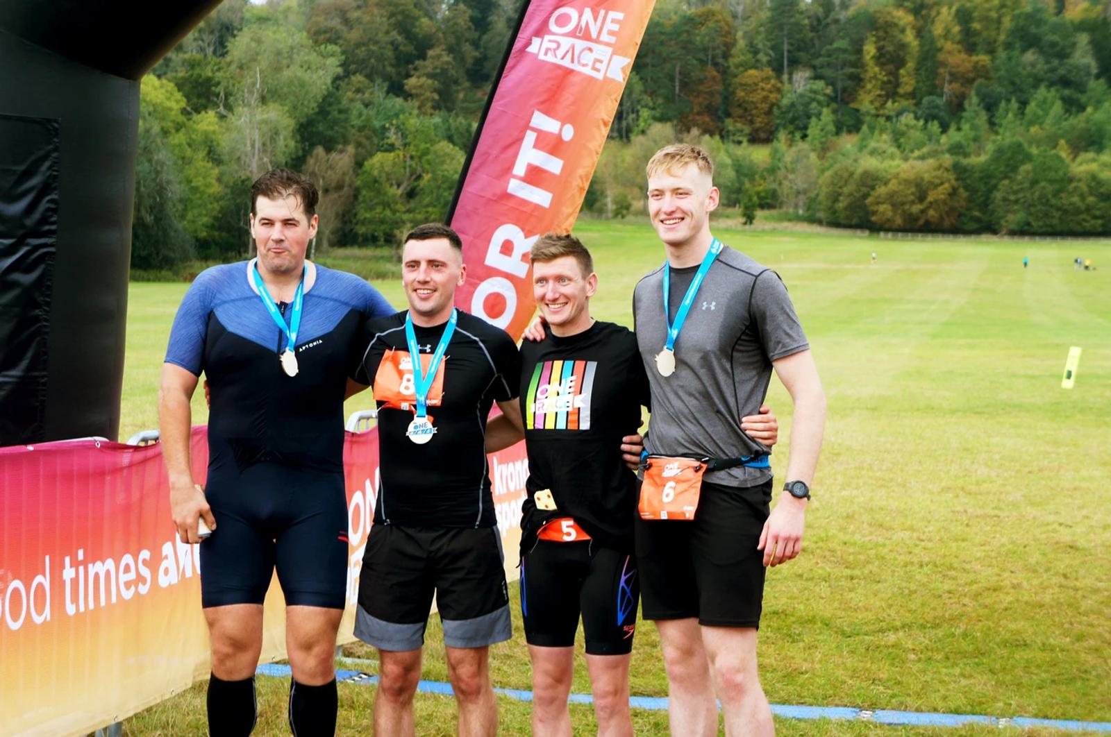 Four men wearing athletic gear and medals stand together smiling in front of a colorful race banner. They're on a grassy field with trees in the background, celebrating after finishing a race.