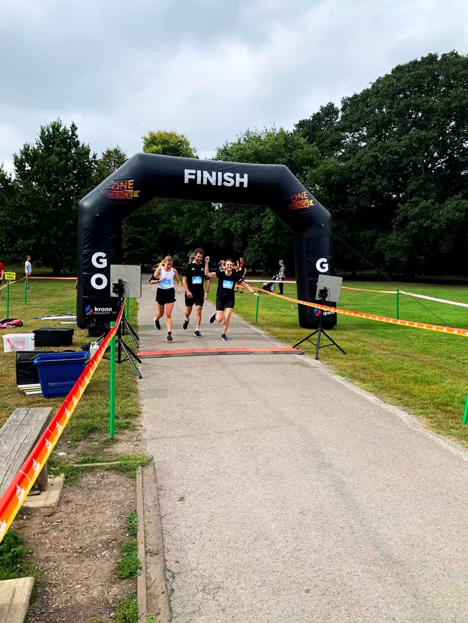 A group of three runners, two women and one man, are celebrating as they cross a race finish line together. The finish line is marked by a black archway under a cloudy sky, and the surrounding area features green grass and trees.