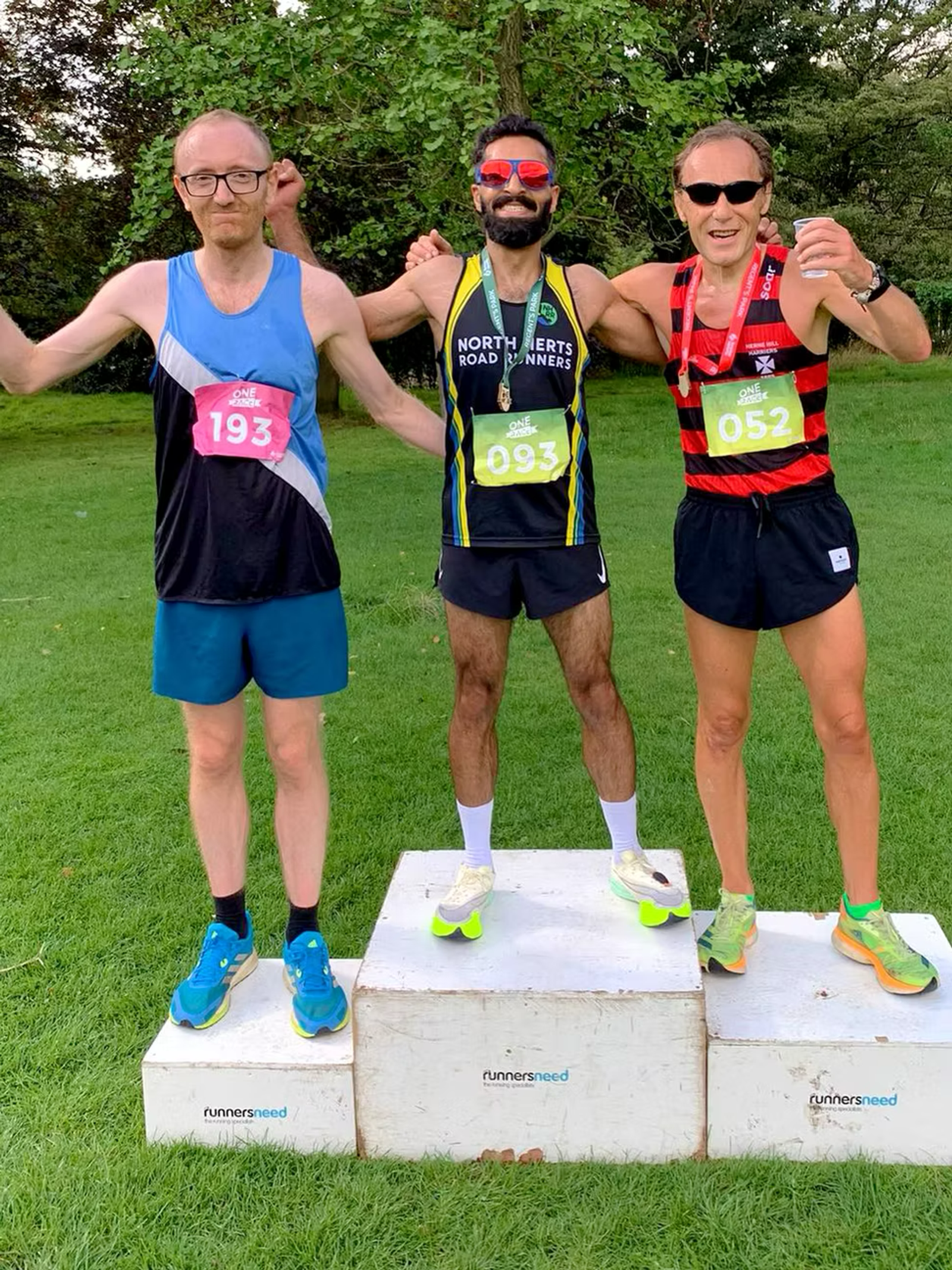 Three men stand on a winners' podium. The middle man is in first place, wearing sunglasses and a black and yellow running jersey. The runner on the right is in second place wearing a red and black jersey, and the left is in third place.
