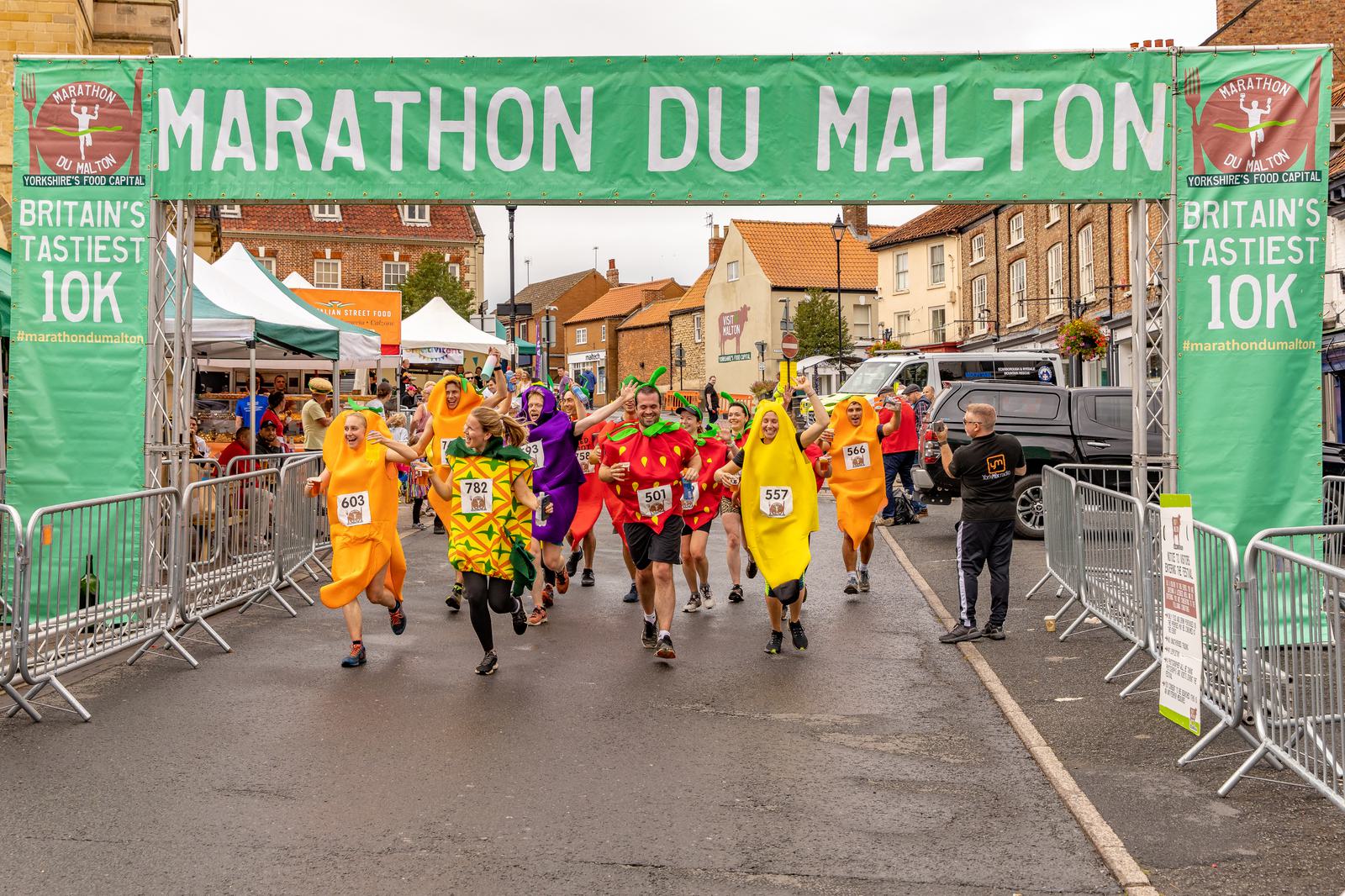 Participants in colorful fruit and vegetable costumes run under a green "Marathon du Malton" banner at the start of a 10K race. The setting is a village street with spectators, tents, and buildings in the background. The event is labeled as "Britain's Tastiest 10K.