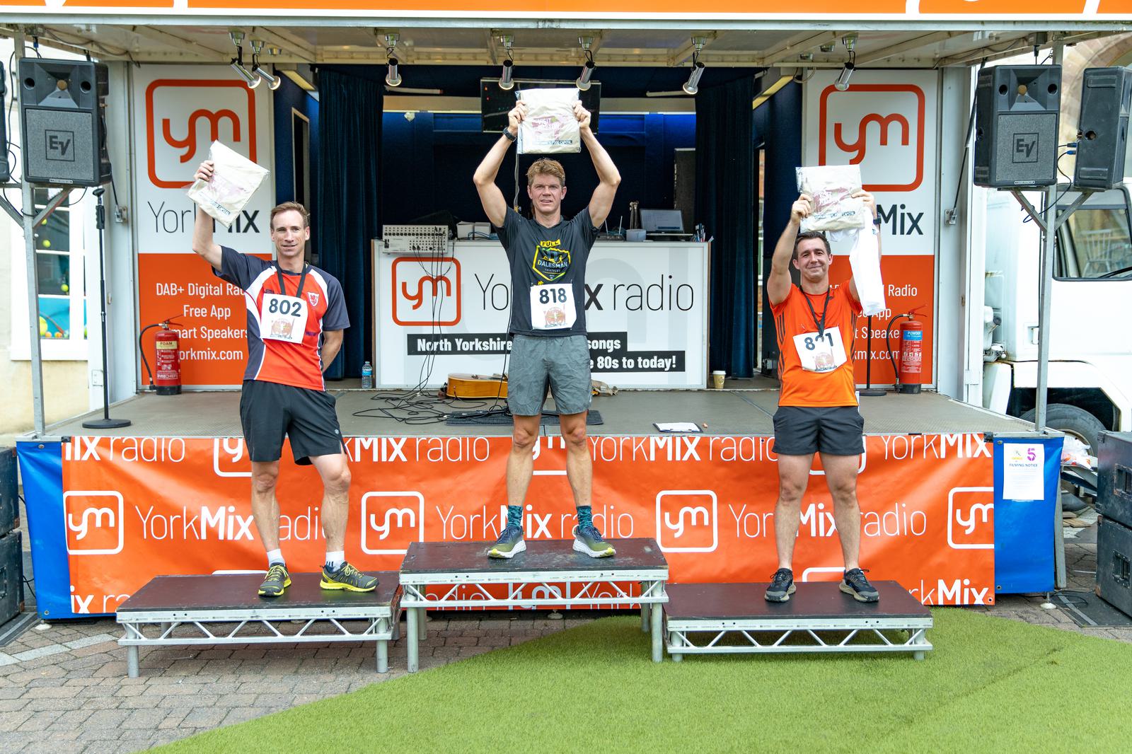 Three male runners stand on podiums holding trophies above their heads. The man in the center is on the highest podium, flanked by two other runners. The event backdrop displays "YorkMix Radio." The runners wear race bibs and medals, indicating their placement.